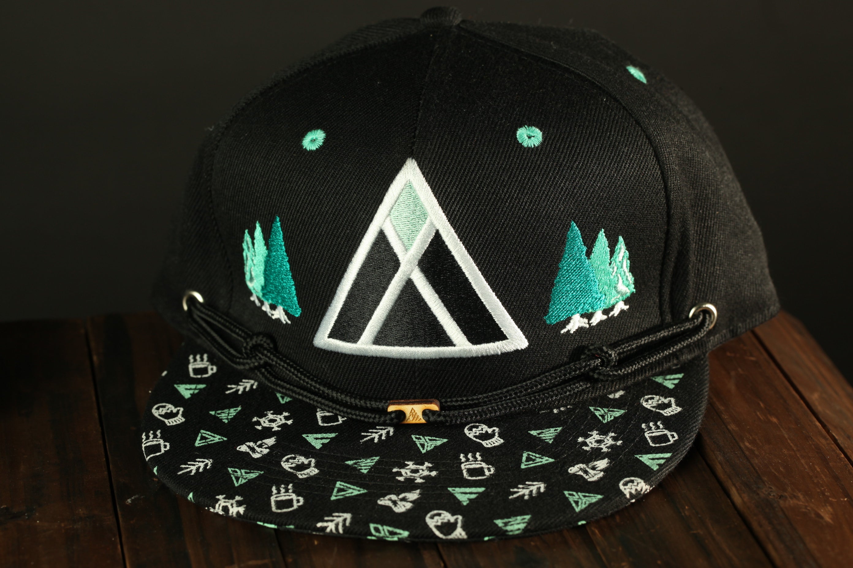 A black baseball cap with a pattern of green and white triangles and trees, along with a small triangle-shaped logo on the front.