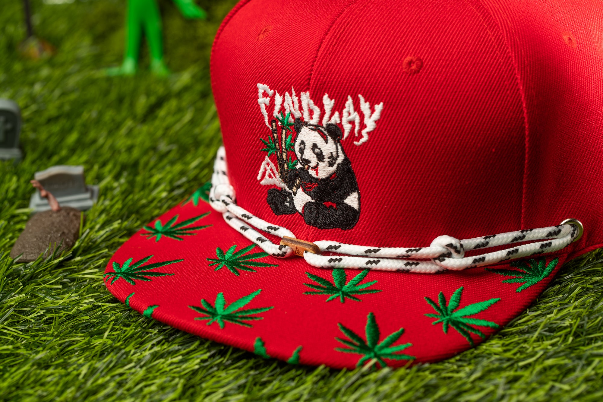 A red baseball cap with a panda bear design and green leaf patterns on the brim, placed on a grassy surface.