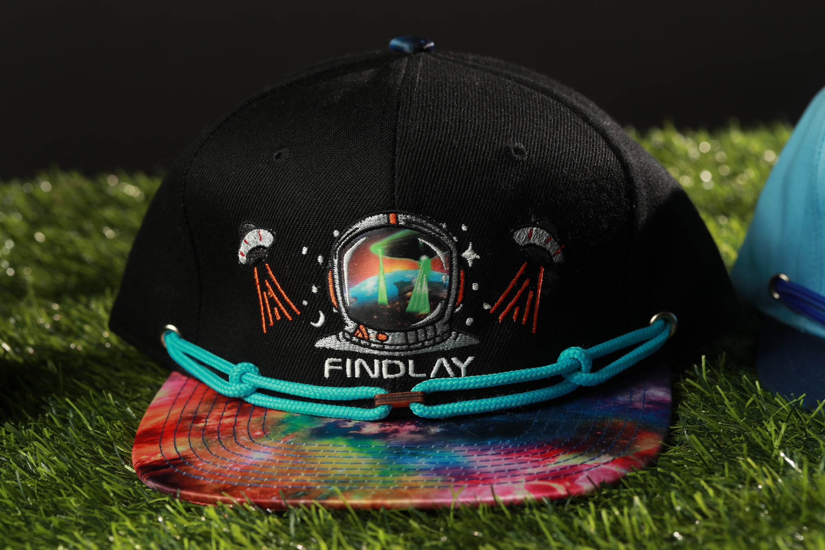 A black baseball cap with a tie-dye pattern and an embroidered image of a space suit, sitting on a grassy surface.