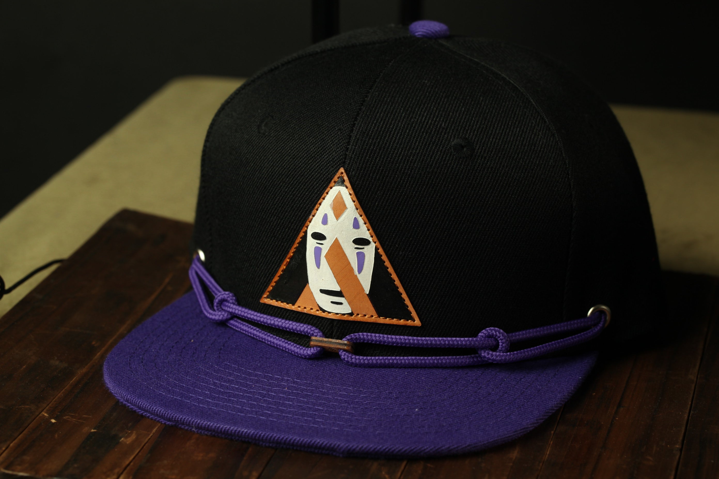 A black baseball cap with a purple brim and a triangular logo on the front, placed on a wooden surface.