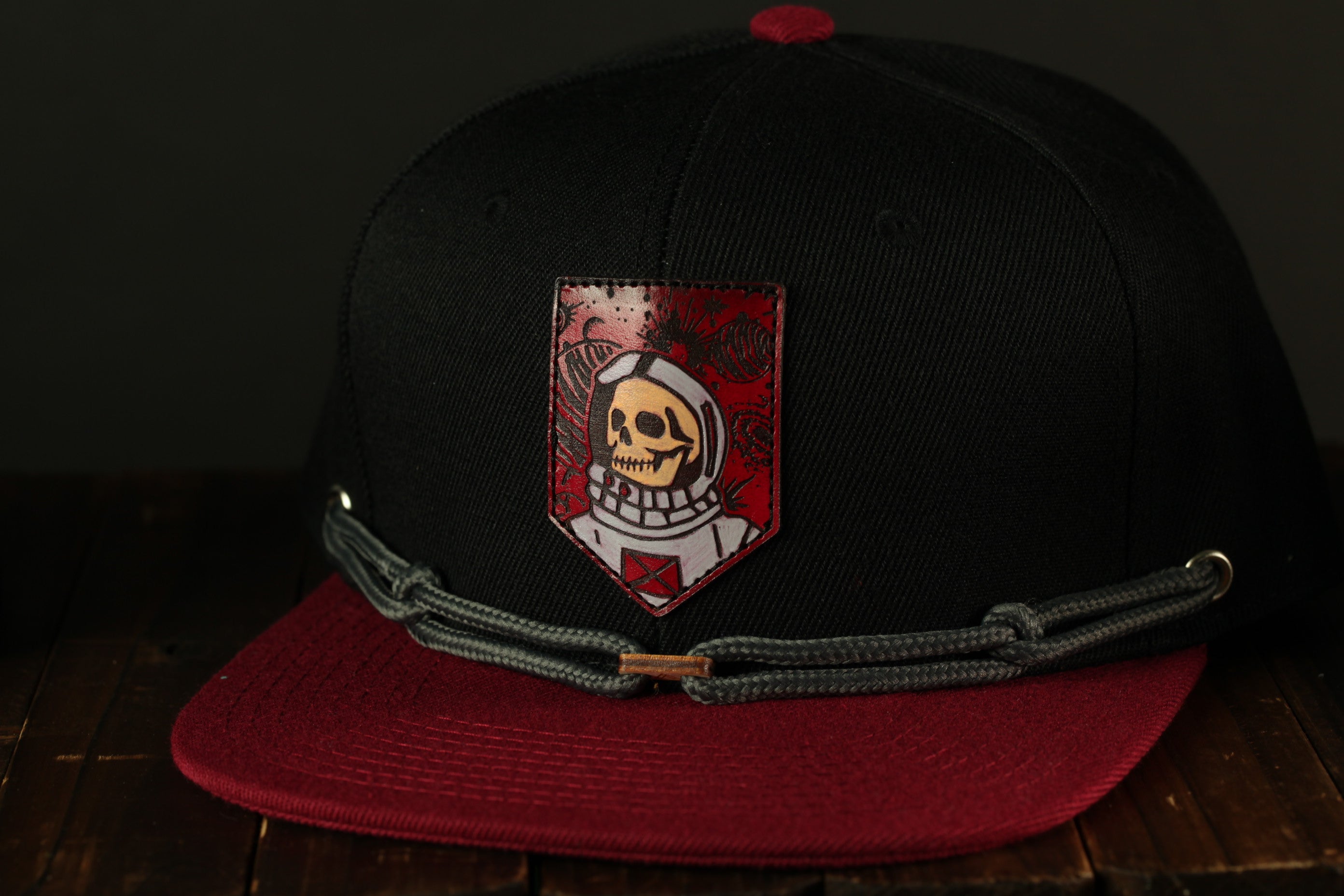 A black baseball cap with a red and gray design featuring a skull wearing a spacesuit, sitting on a wooden surface.