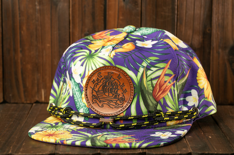 A colorful, patterned baseball cap with a leather patch featuring a ship design, placed on a wooden surface.