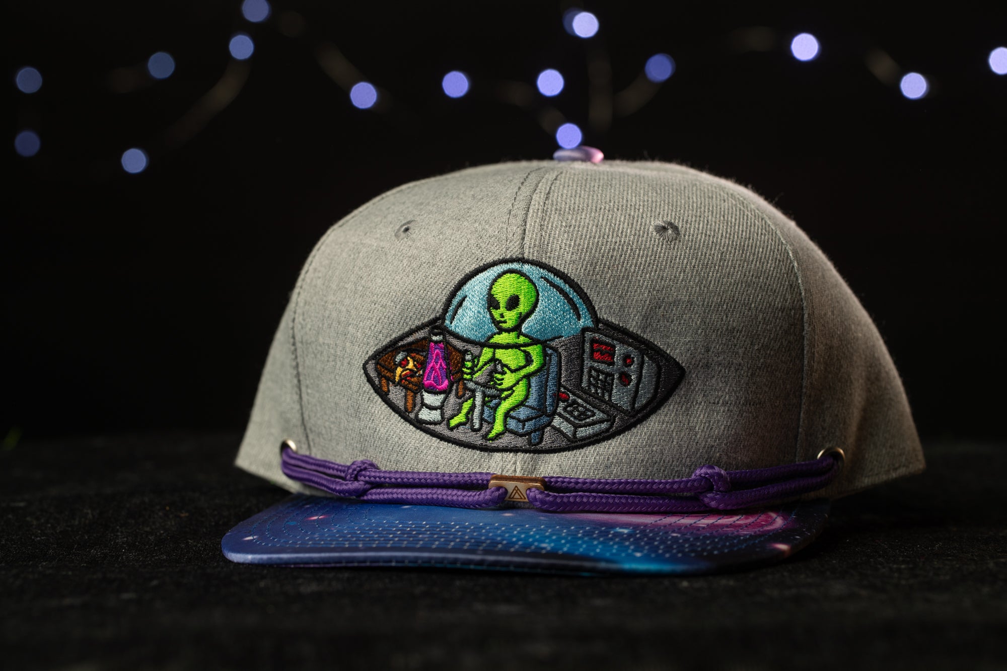 A gray baseball cap with a colorful alien embroidered on it, set against a dark background with twinkling lights.