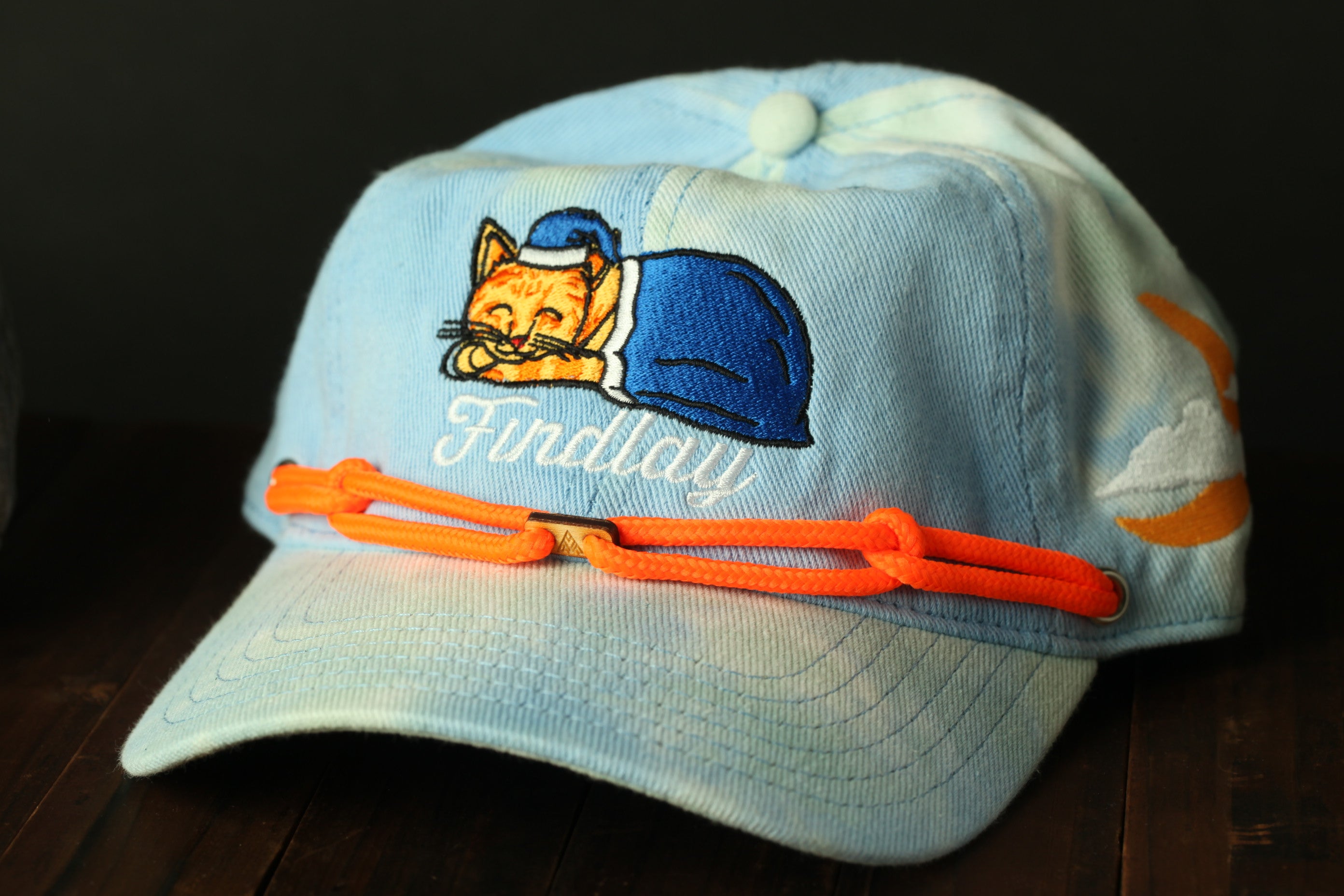 A blue baseball cap with a cat embroidered on it, featuring a sleeping cat and the word "Findlay" written on it.