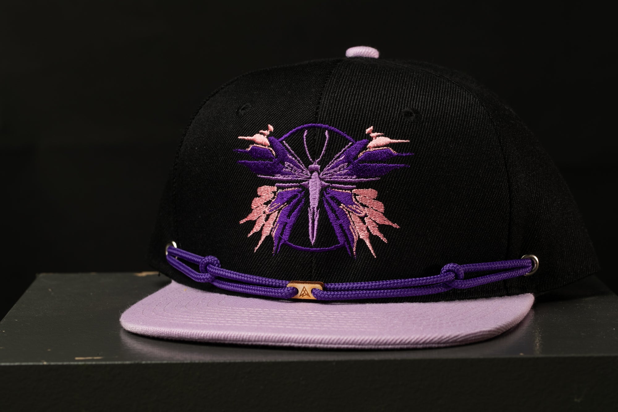 A black baseball cap with a purple and pink butterfly embroidered on it, sitting on a gray surface.