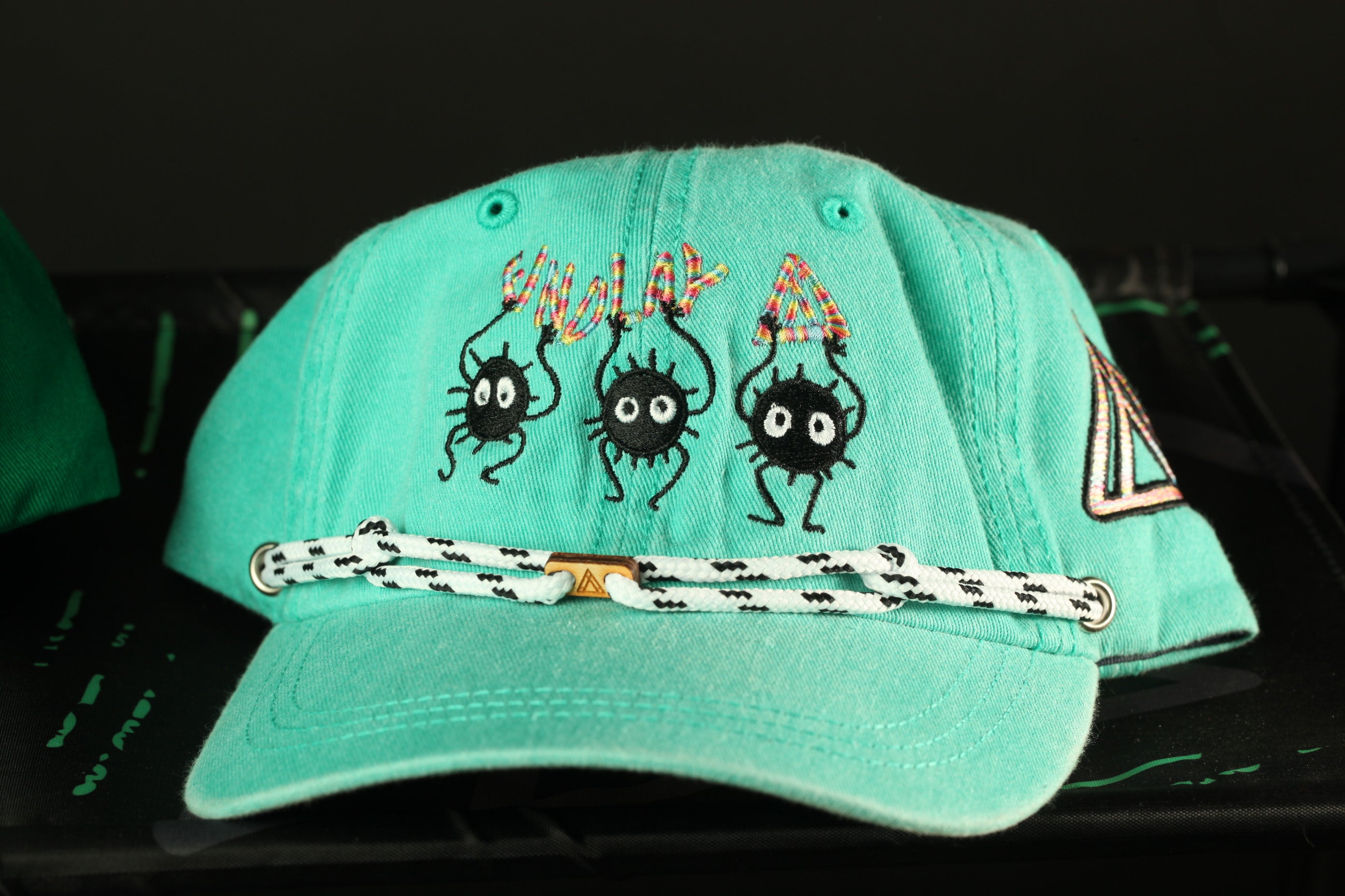 A turquoise baseball cap with a white and black braided strap, featuring three cartoon-like black creatures with large eyes and legs.
