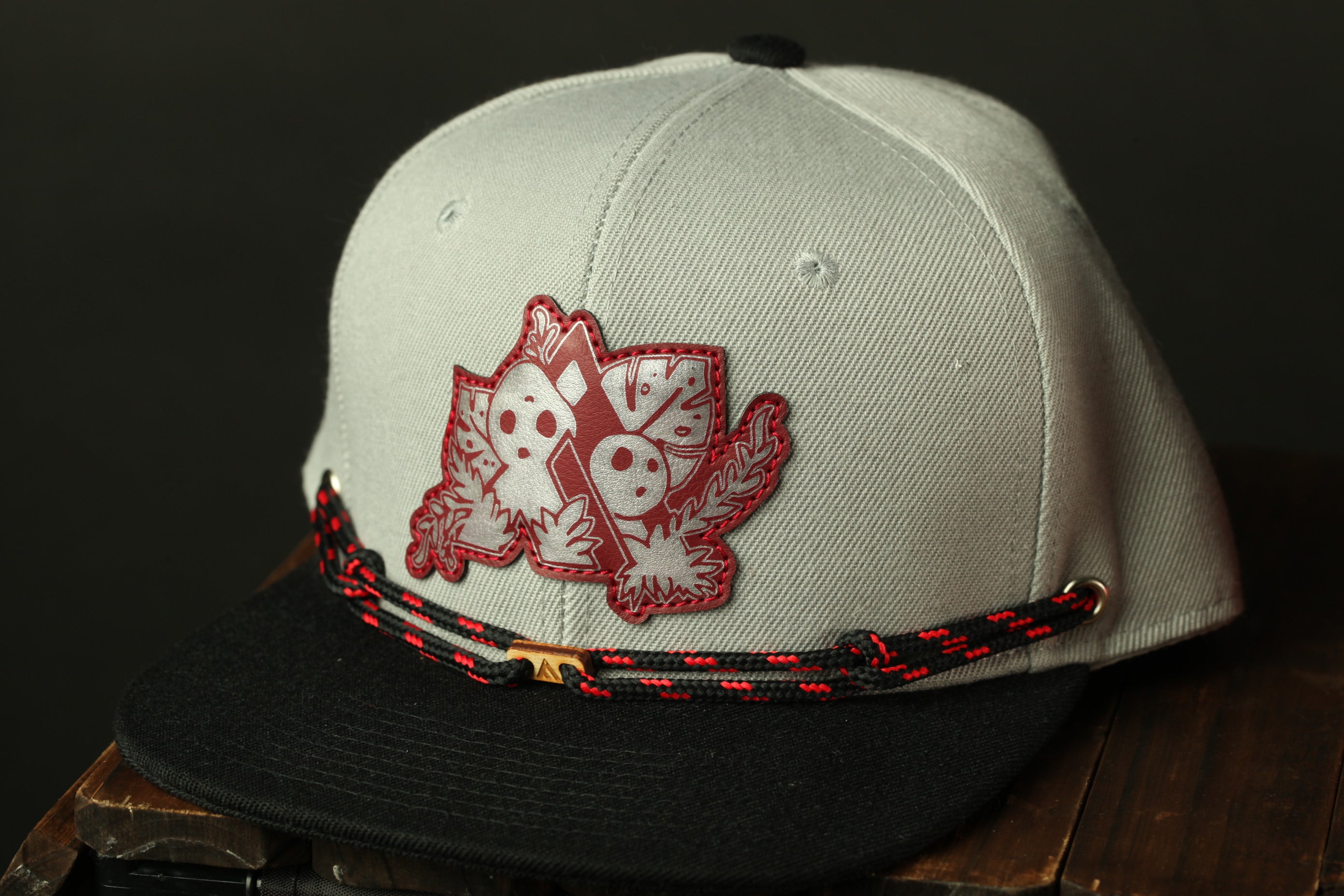 A gray baseball cap with a red and black braided strap and a patch featuring a stylized design on the front.