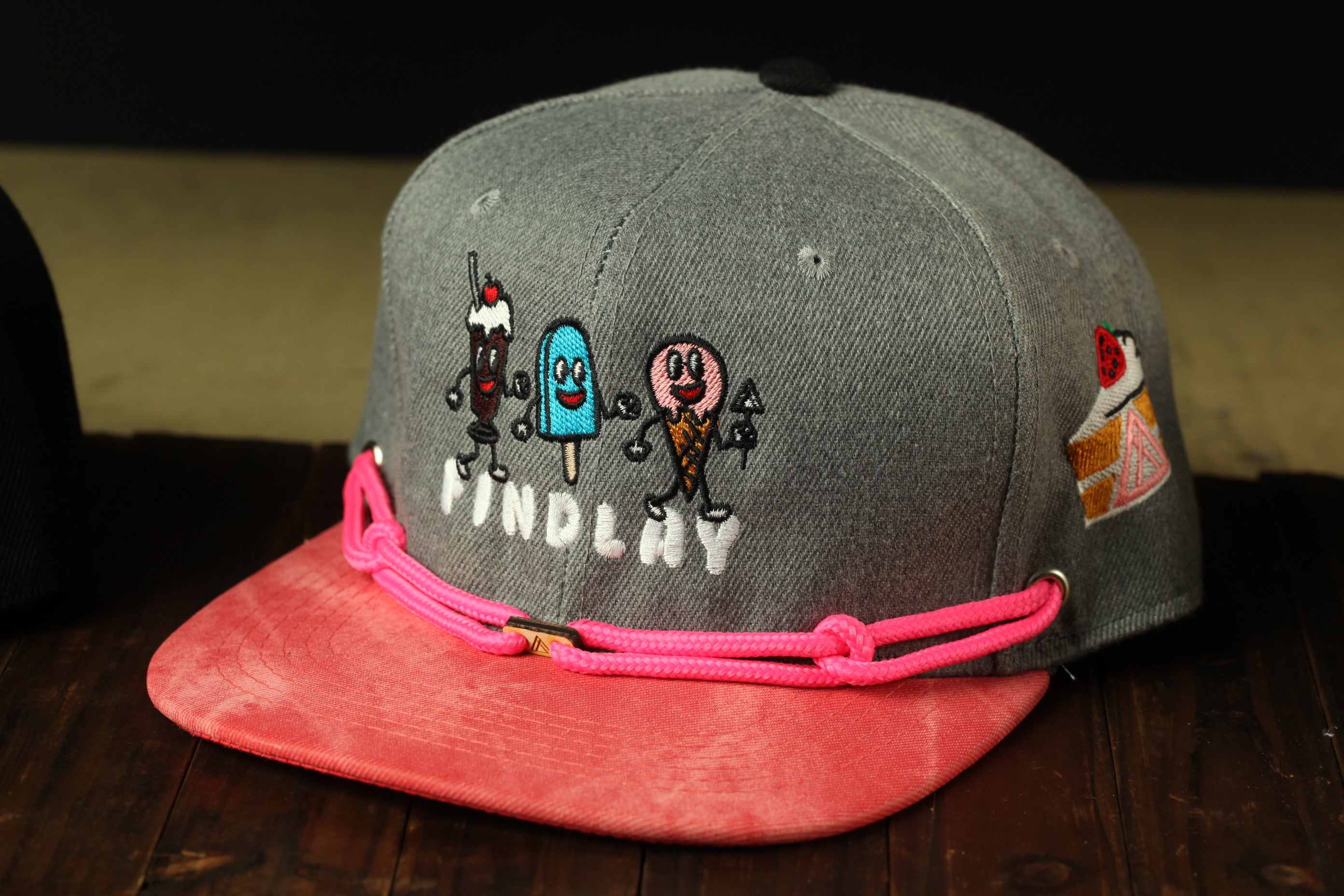 A gray baseball cap with a pink strap and cartoon characters on it, sitting on a wooden surface.