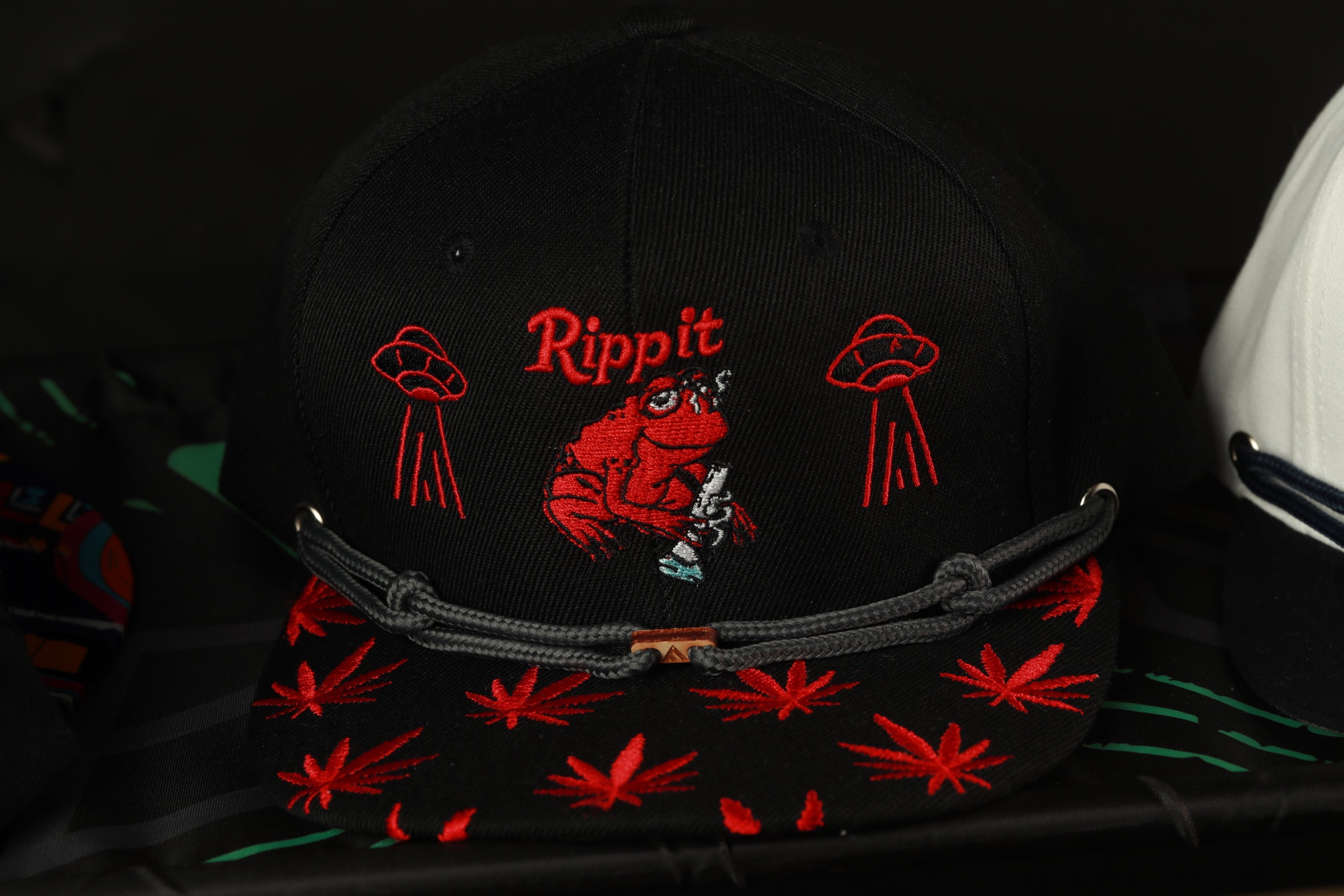 A black baseball cap with red embroidered designs, including a red frog and two red alien-like figures, and the word "Rippit" printed on it.
