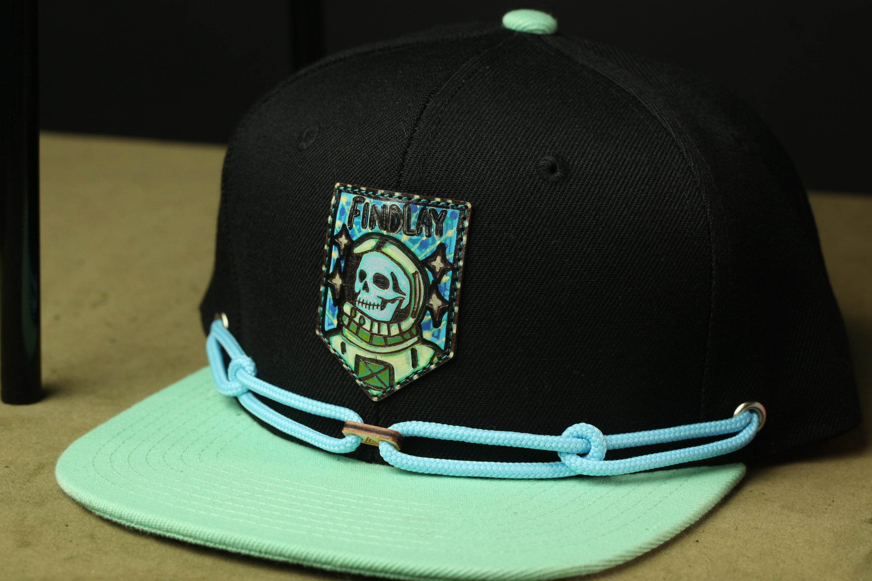 A black baseball cap with a skull and crossbones design, a blue chain strap, and a small patch on the front.