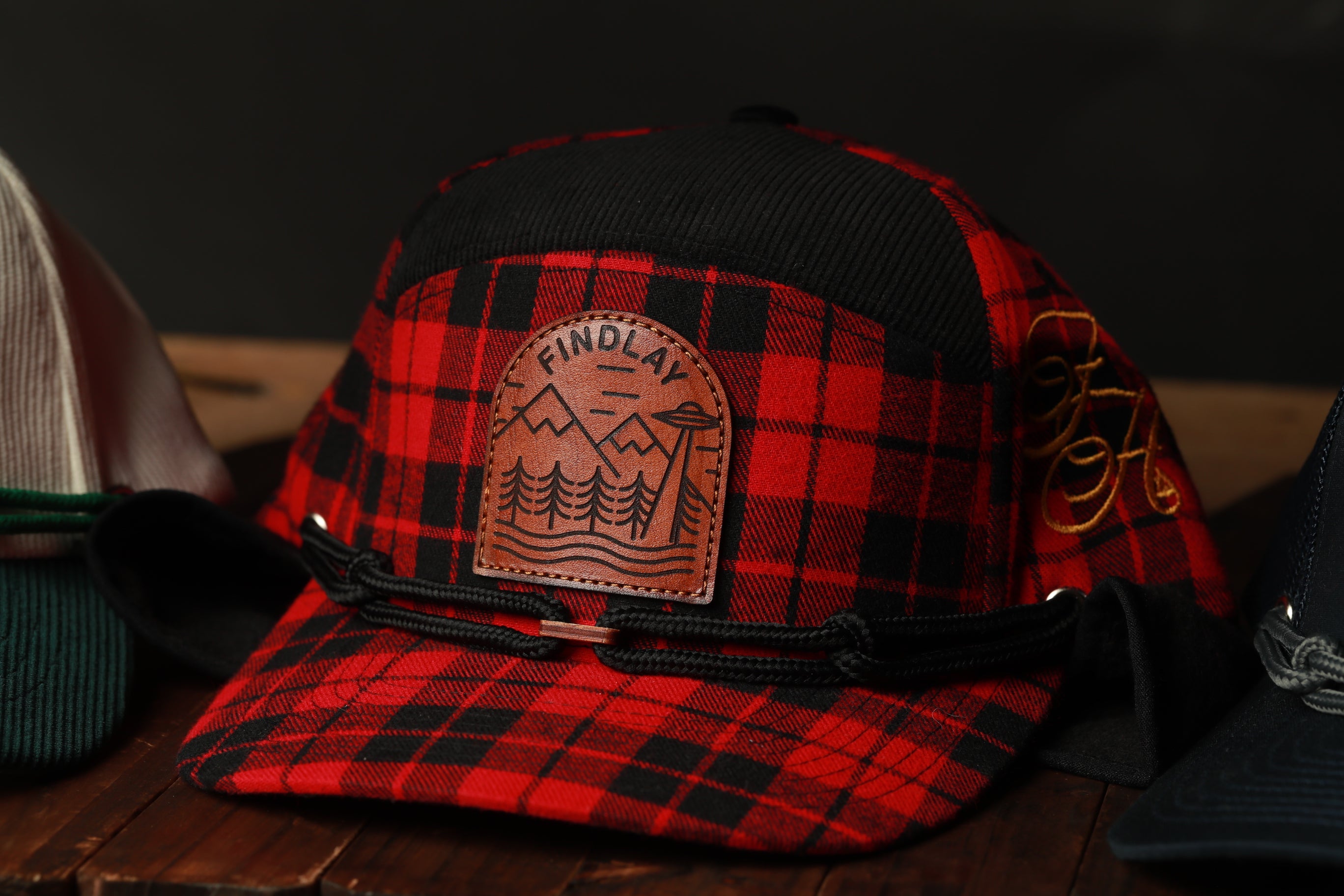 A red and black plaid baseball cap with a leather patch featuring a mountain landscape and the word "FINDLAY" on it.