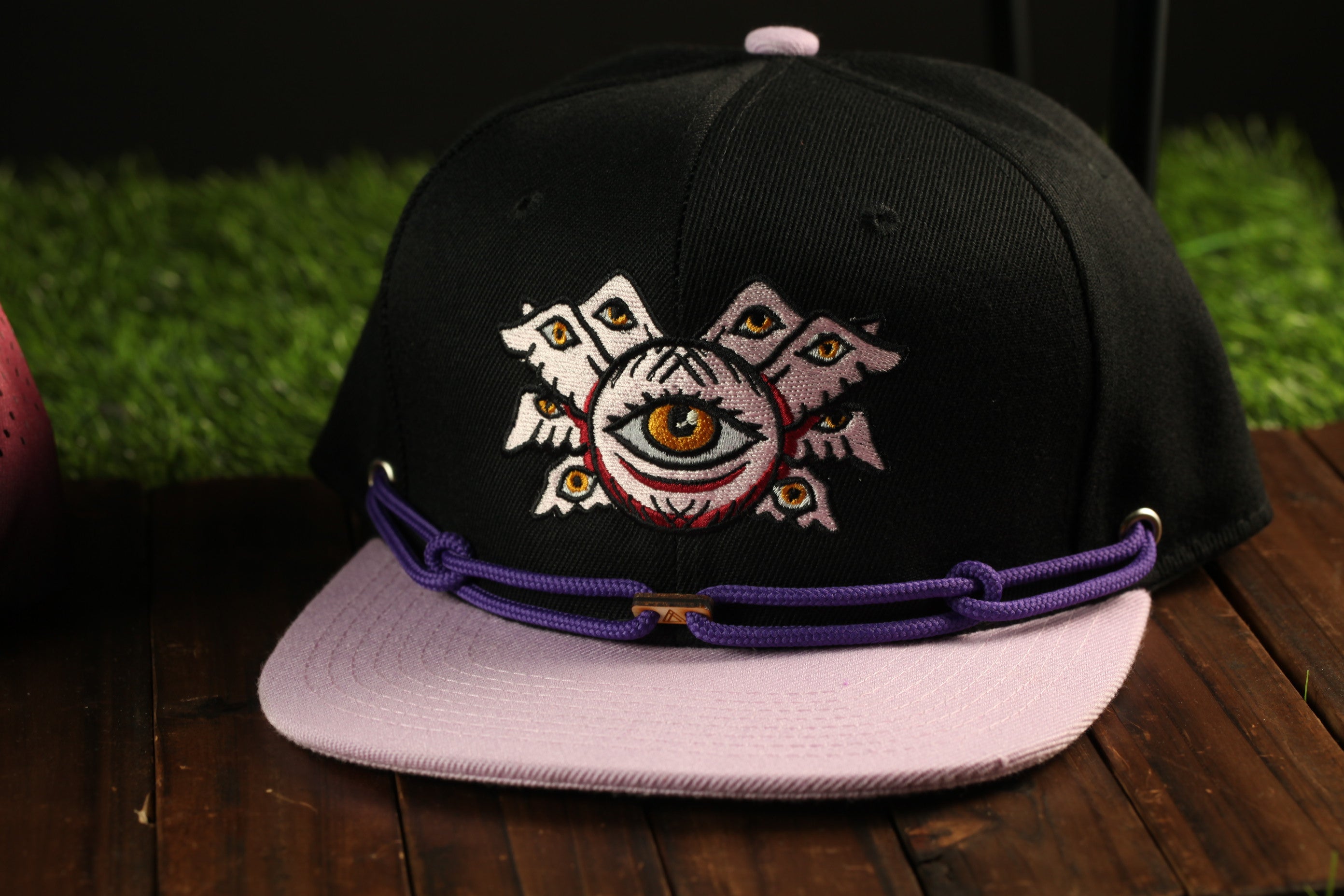 A black baseball cap with a purple band and an eye design on the front, placed on a wooden surface.