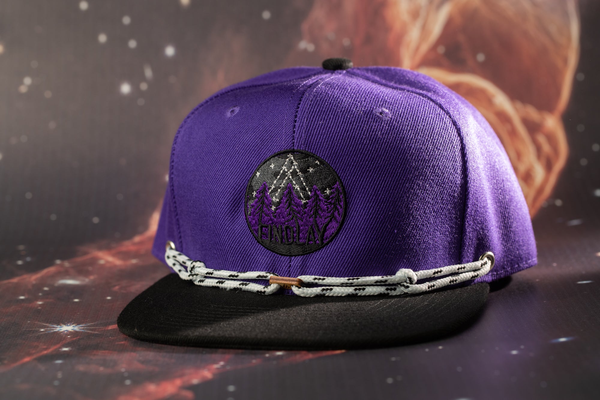 A purple baseball cap with a black brim and a white lace strap, featuring a circular logo with a mountain and tree design.