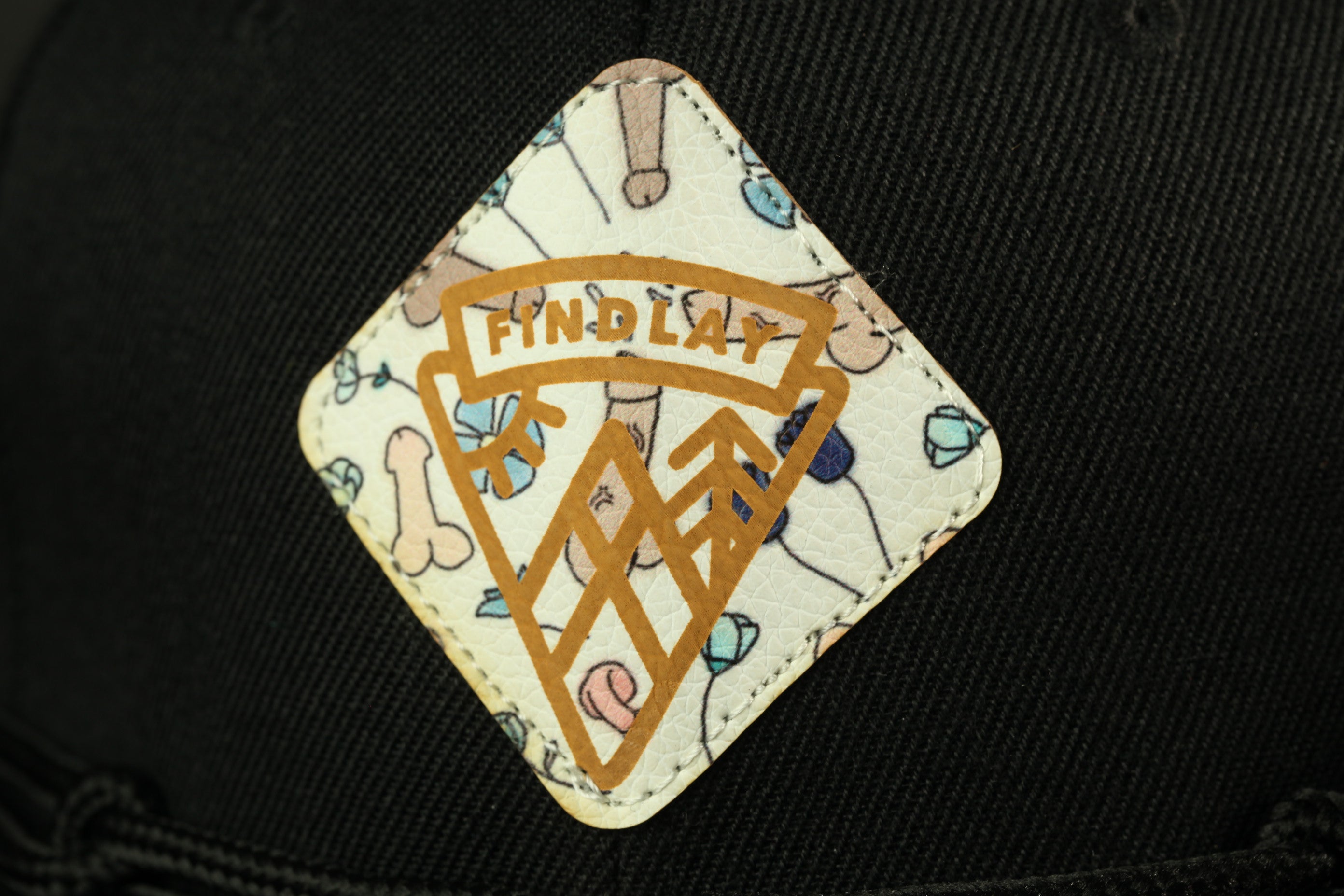 A close-up of a black baseball cap with a white and gold patterned patch featuring a logo.