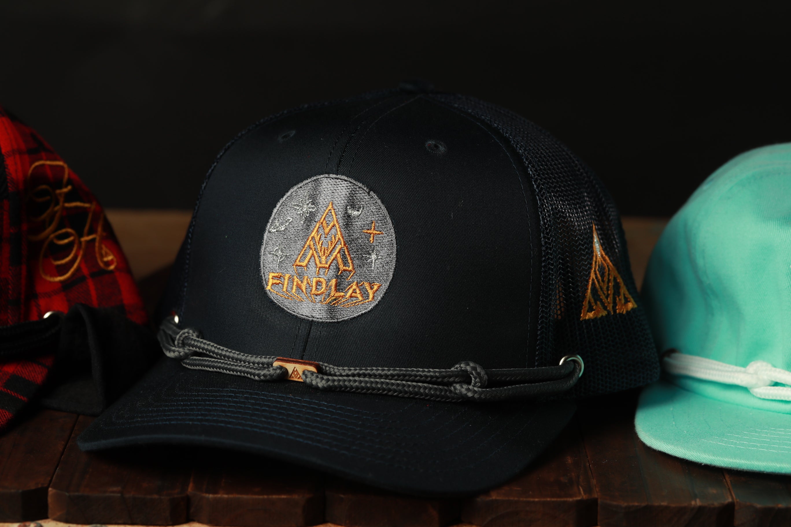 A black baseball cap with a logo on it, sitting on a wooden surface next to a teal baseball cap.