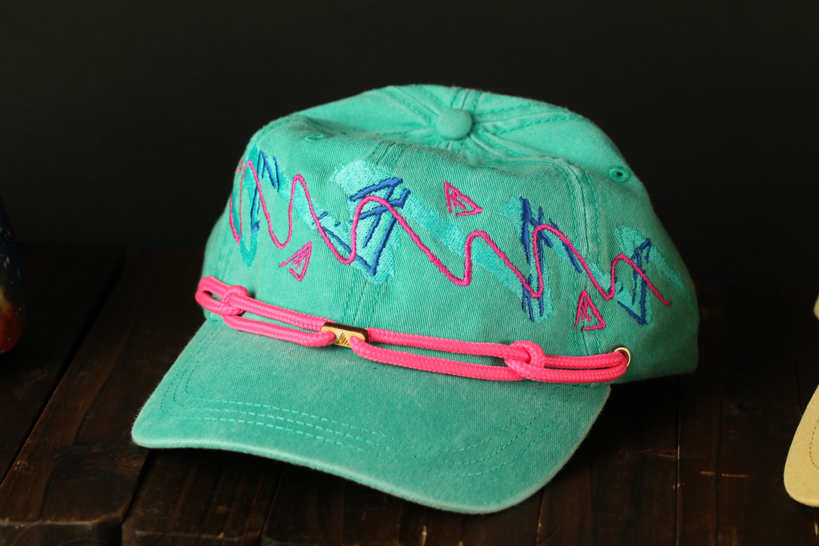 A turquoise baseball cap with pink braided straps and colorful geometric designs on the front.