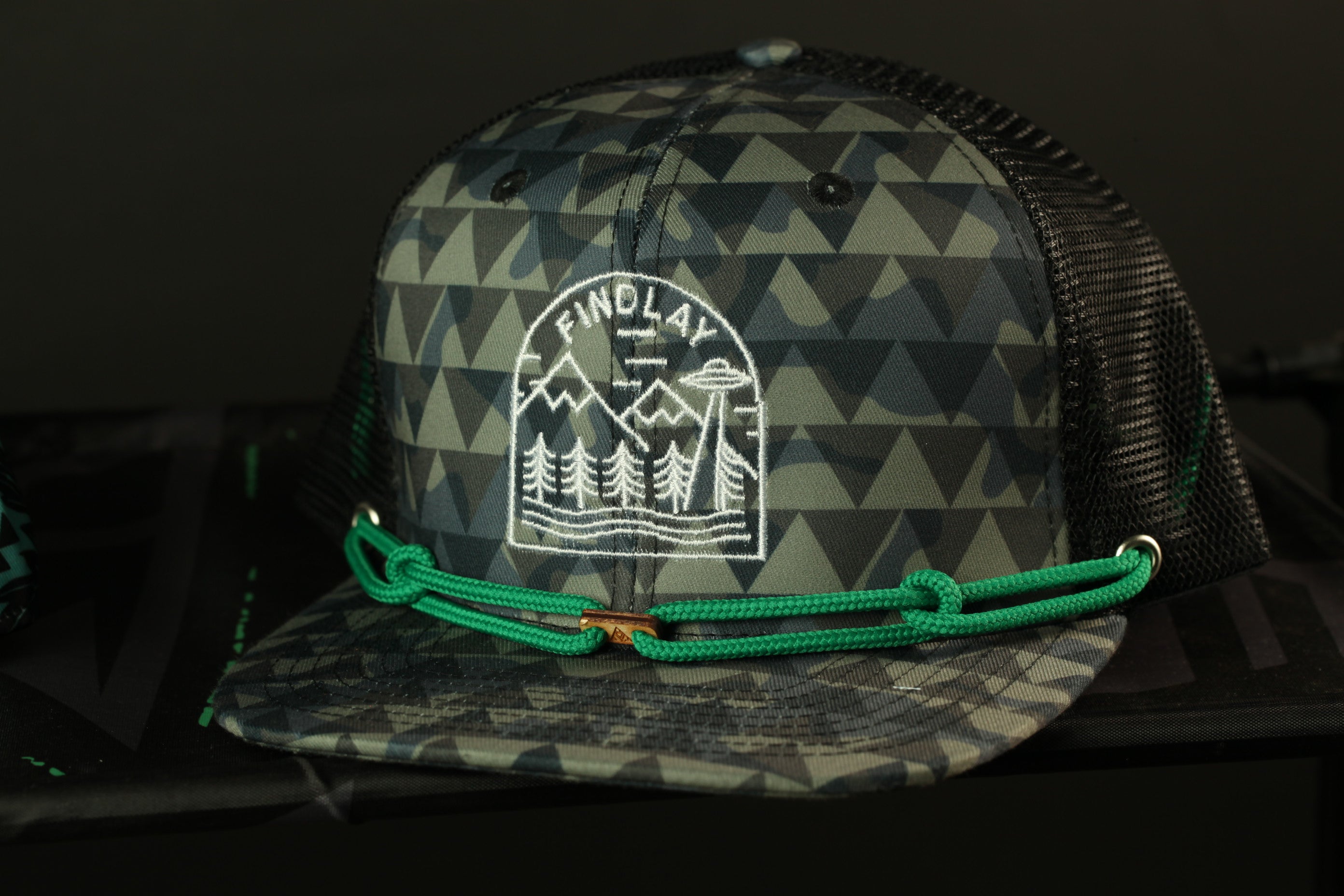 A black and gray camouflage baseball cap with a green rope strap and a white embroidered logo on the front.