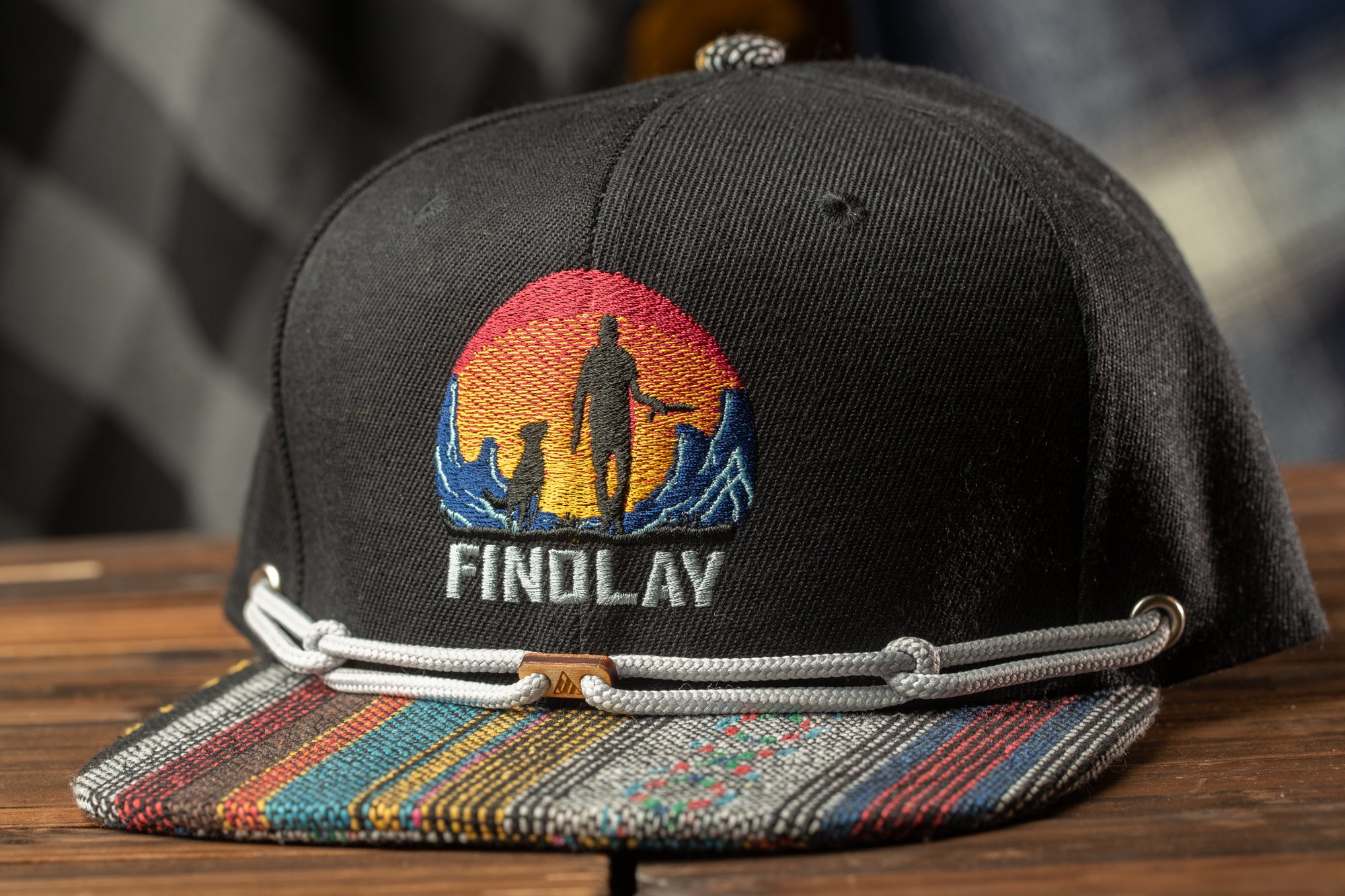 A black baseball cap with a colorful, embroidered design on the front, featuring a silhouette of a person and mountains.