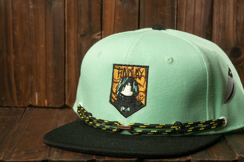 A light green baseball cap with a black brim and a yellow and black lacing pattern, featuring a patch with a cartoon character and the word "FRIDAY" printed on it.