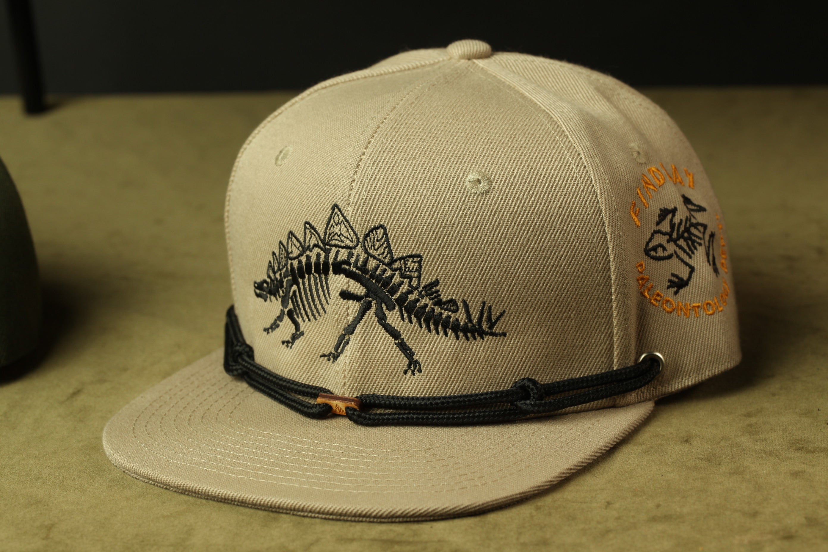 A beige baseball cap with a dinosaur design on the front, featuring a black strap and a small orange logo on the side.