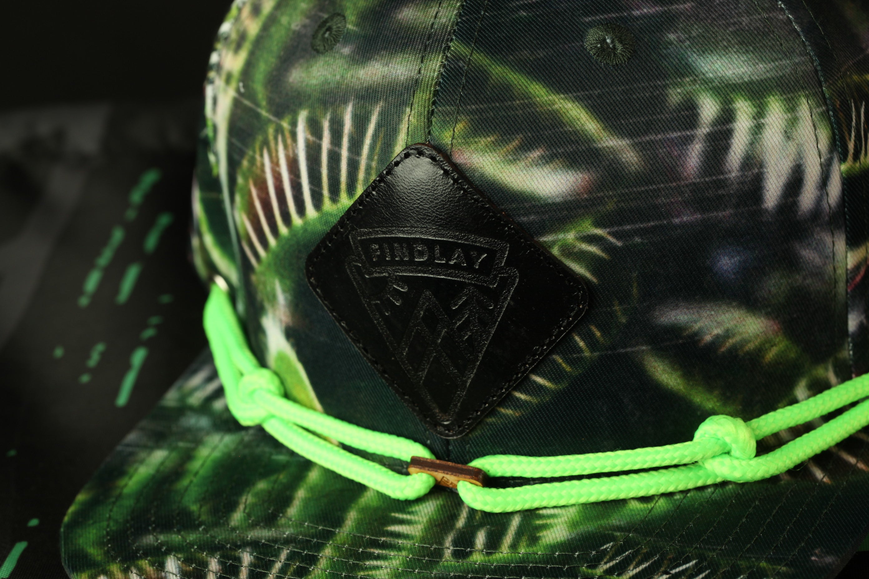 A close-up view of a green and black camouflage-patterned baseball cap with a black leather patch on the front.