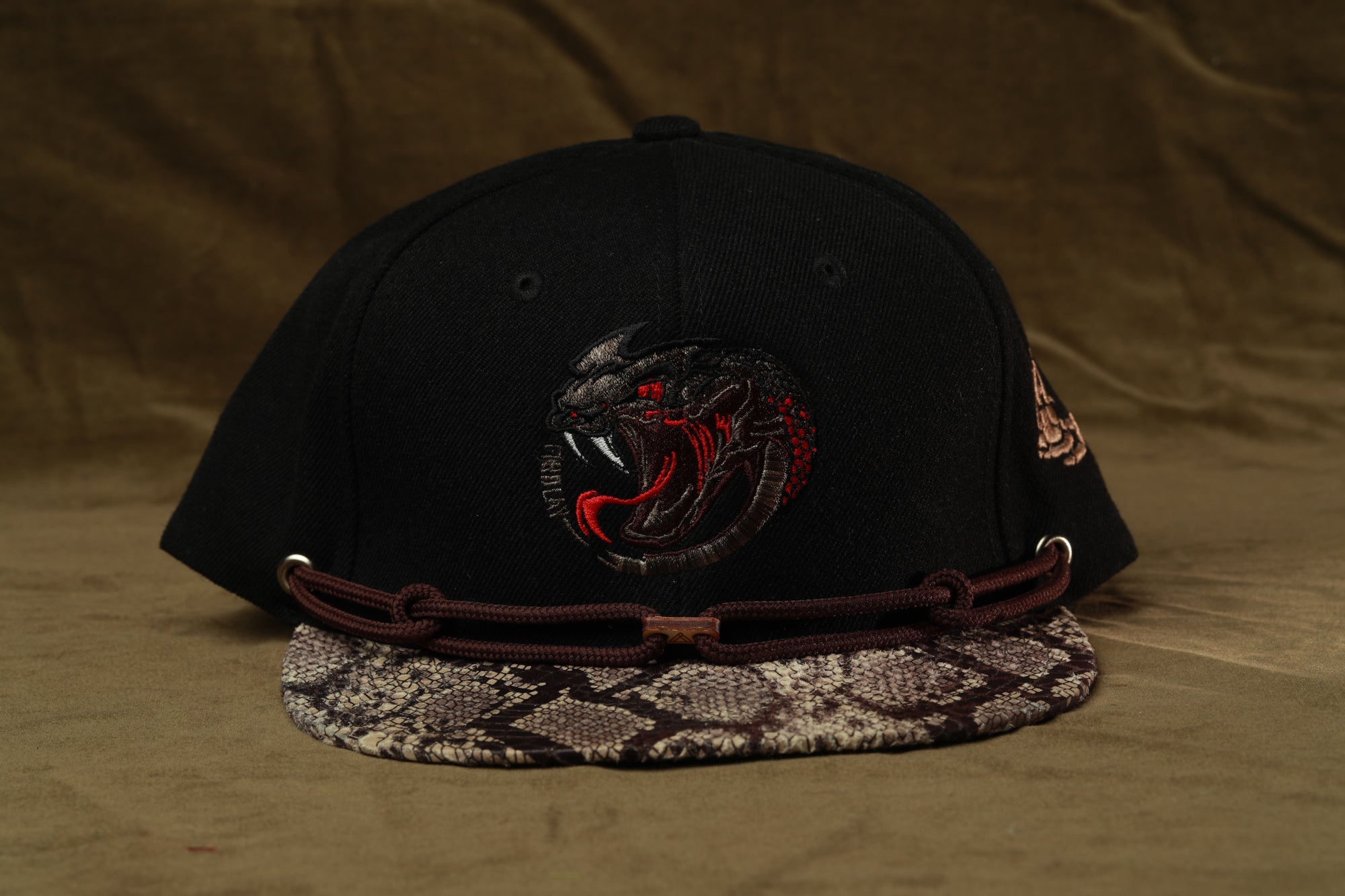 A black baseball cap with a red and black snake design on the front, placed on a brown fabric background.