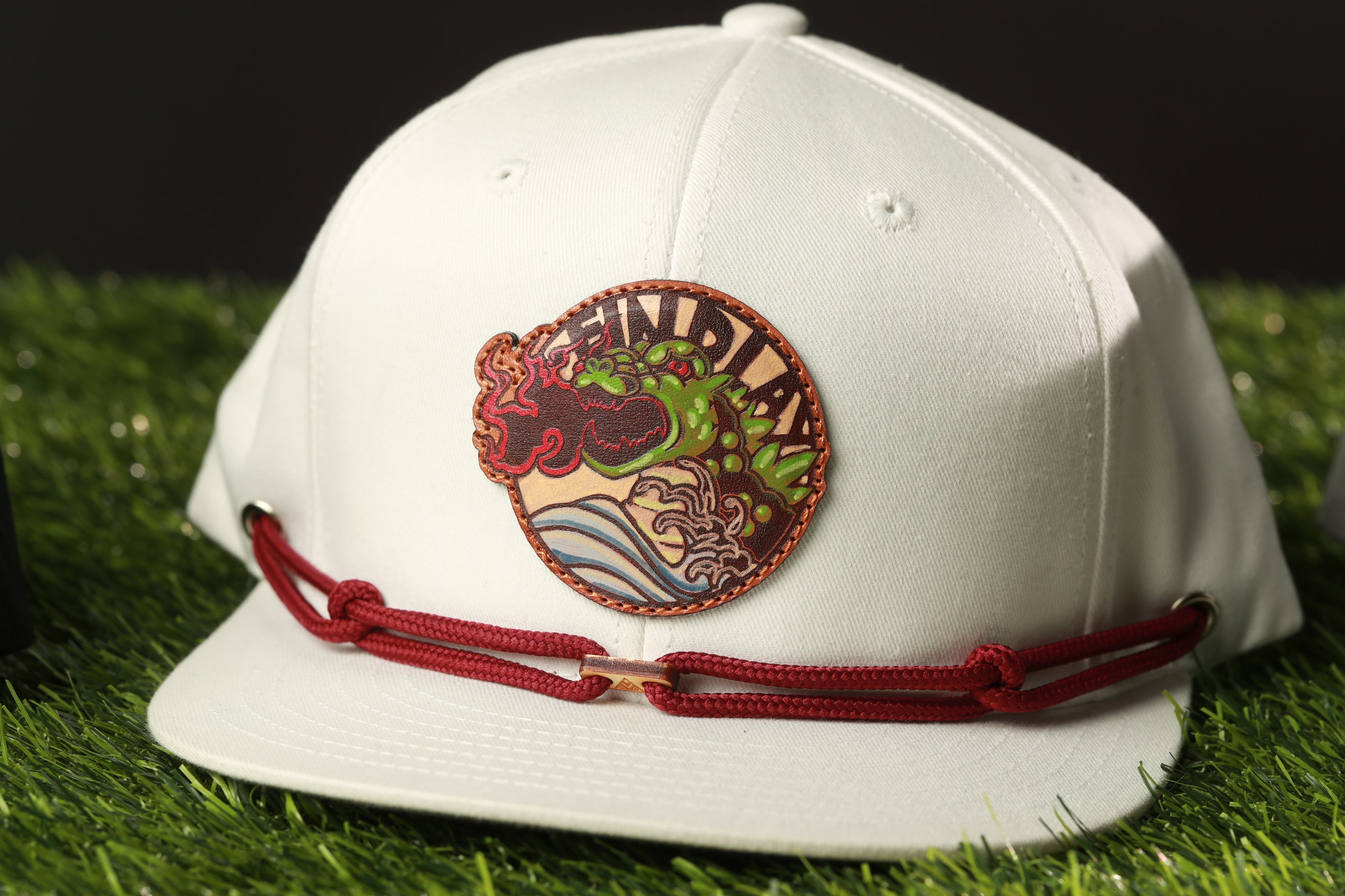 A white baseball cap with a red and brown circular patch featuring a dragon design, placed on a grassy surface.