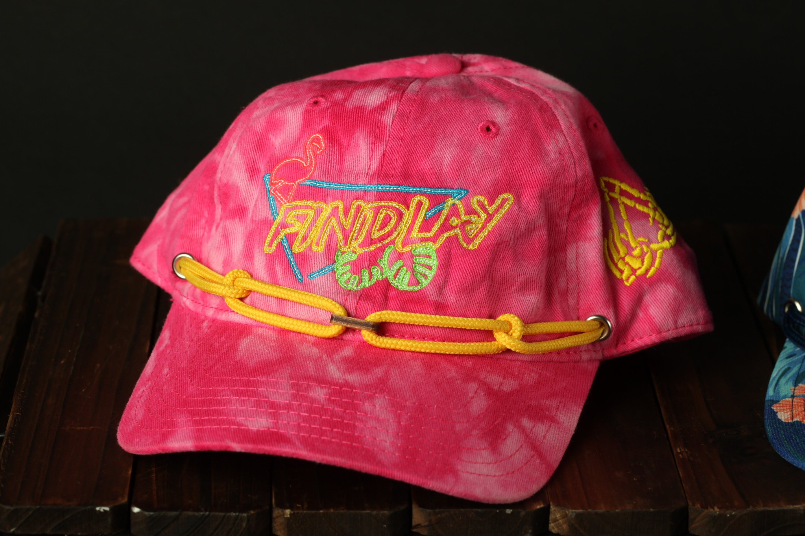 A pink baseball cap with a chain strap and the word "FIND PLAY" embroidered on it, sitting on a wooden surface.