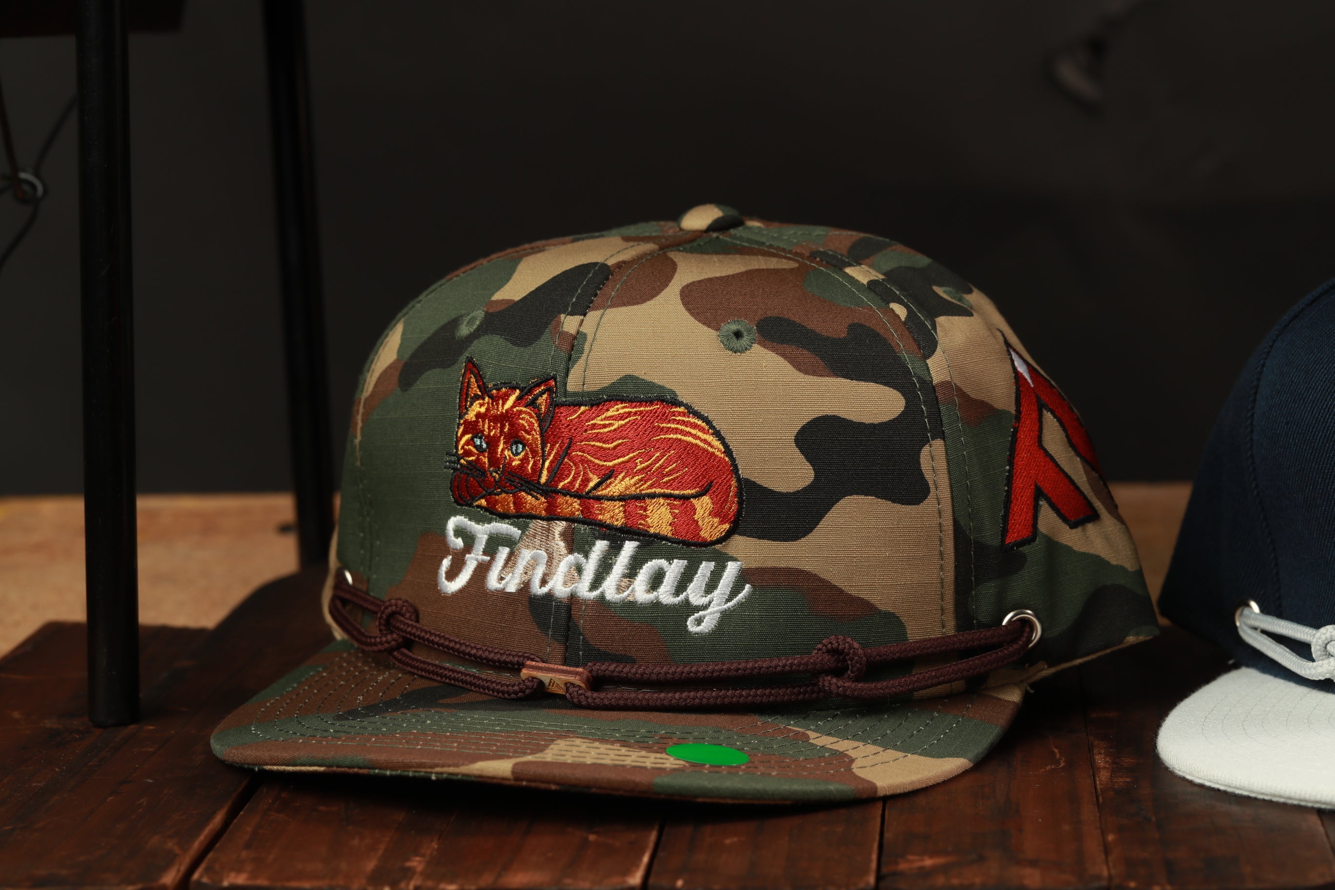 A camouflage baseball cap with a cat embroidered on it, sitting on a wooden surface.