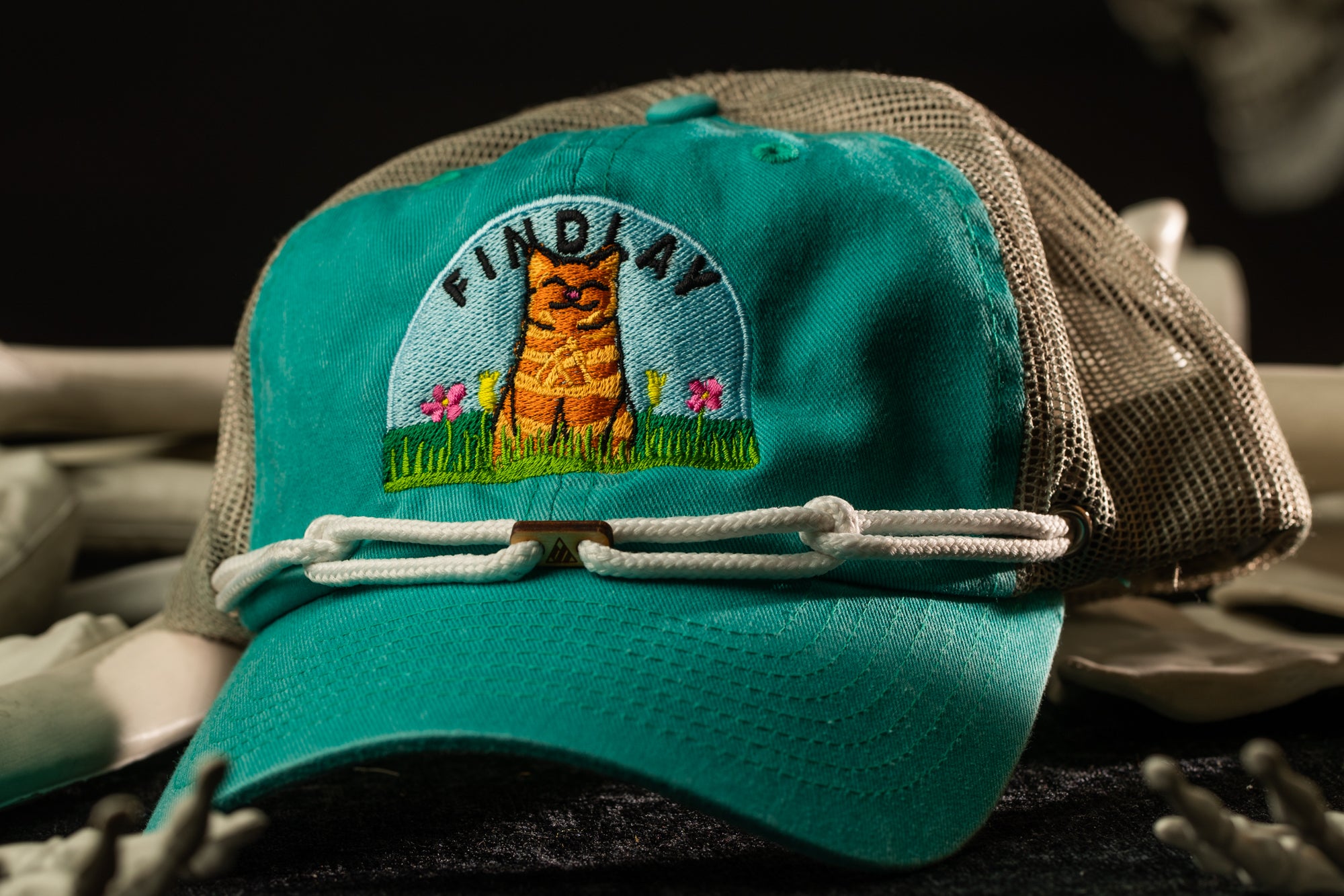 A turquoise baseball cap with a cat embroidered on it, sitting on a surface with a mesh pattern.