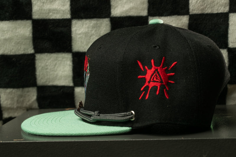 A black baseball cap with a red triangle design and a green brim, placed on a black surface against a checkered background.