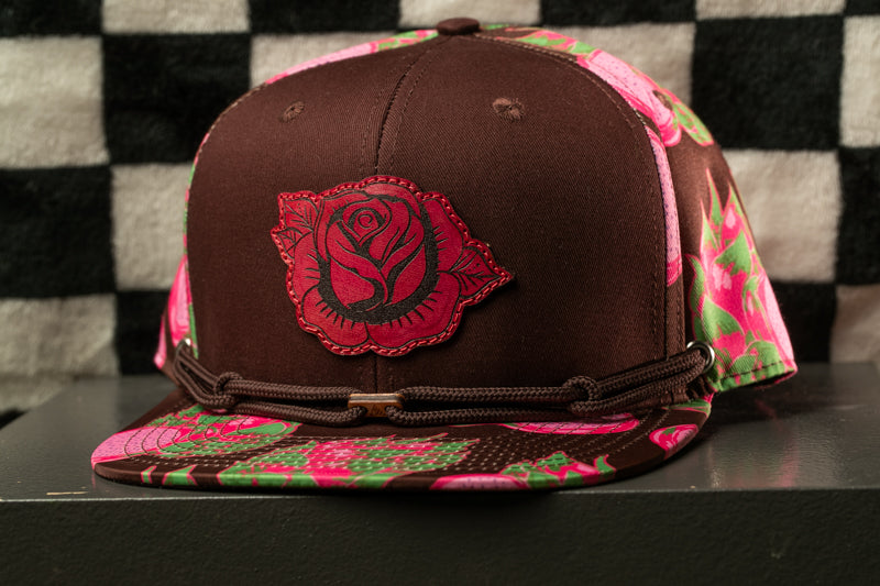 A brown baseball cap with a red rose design on the front, placed on a black surface against a checkered background.