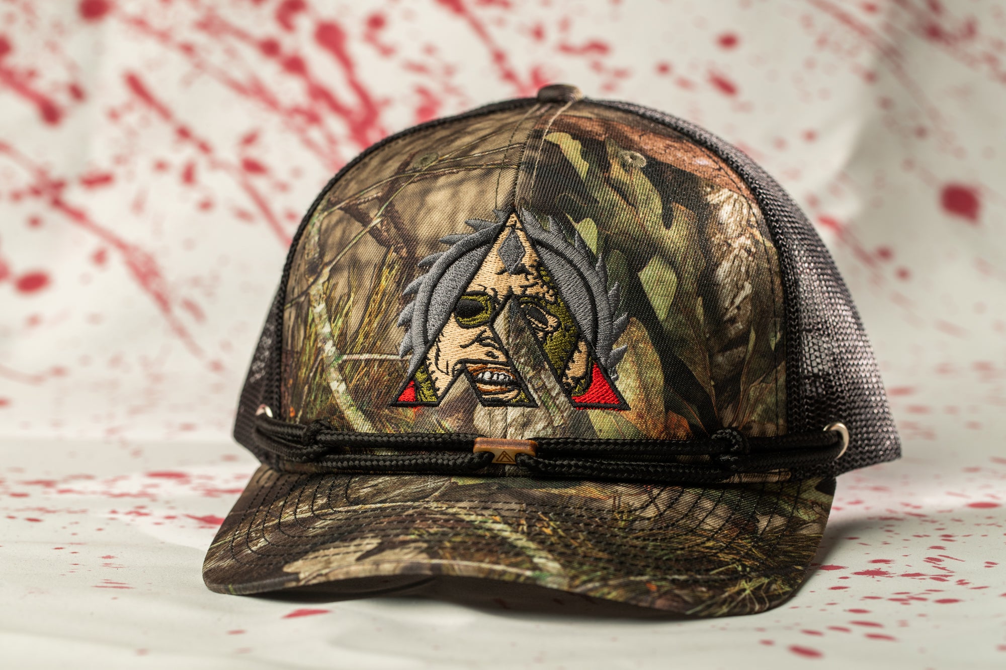 A camouflage baseball cap with a skull embroidered on it, set against a background of red splatters.
