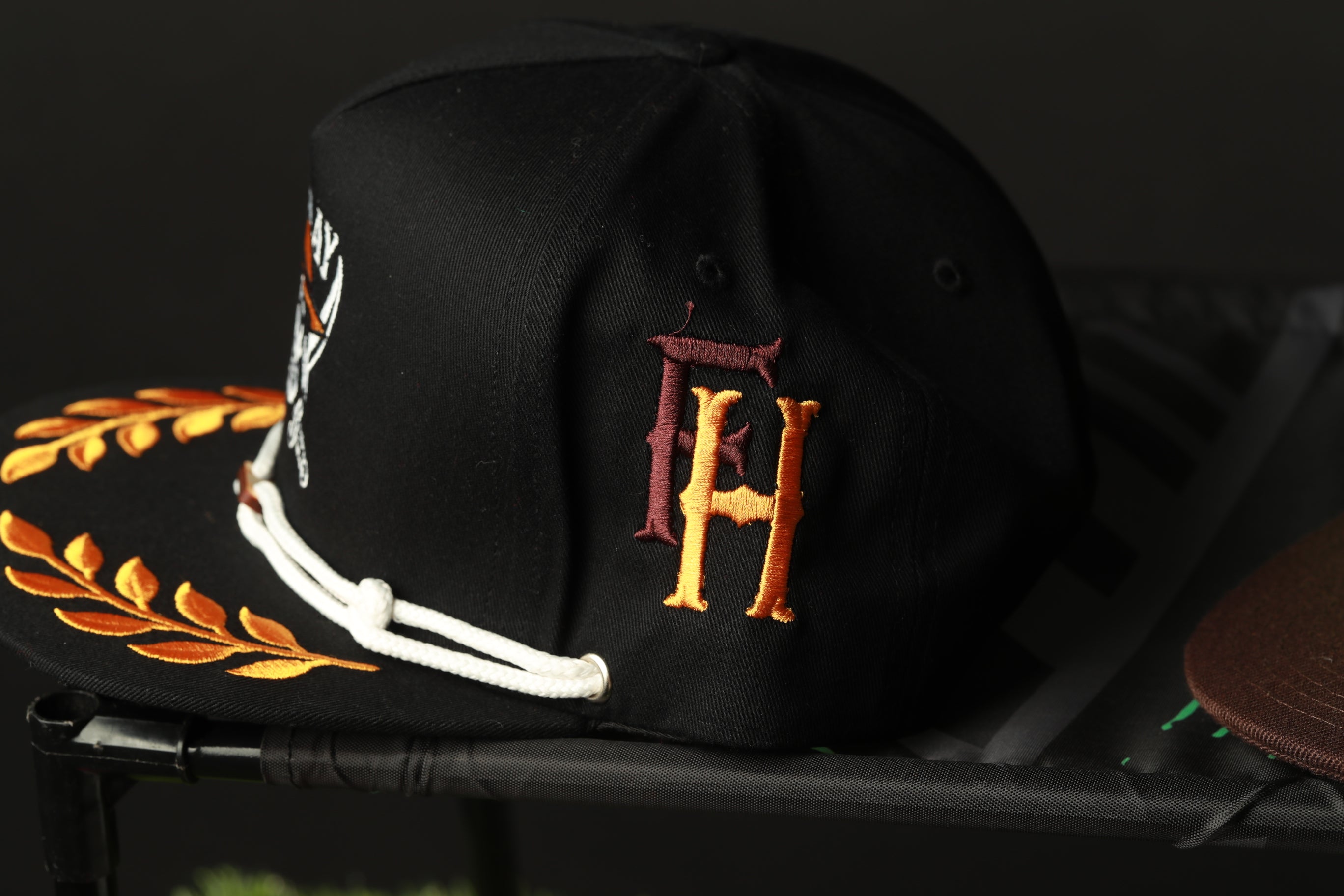 A black baseball cap with a gold and maroon embroidered logo on the side, sitting on a black surface.