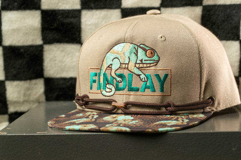A beige baseball cap with a graphic of a chameleon and the word "FINDlay" printed on it, sitting on a surface with a checkered pattern in the background.