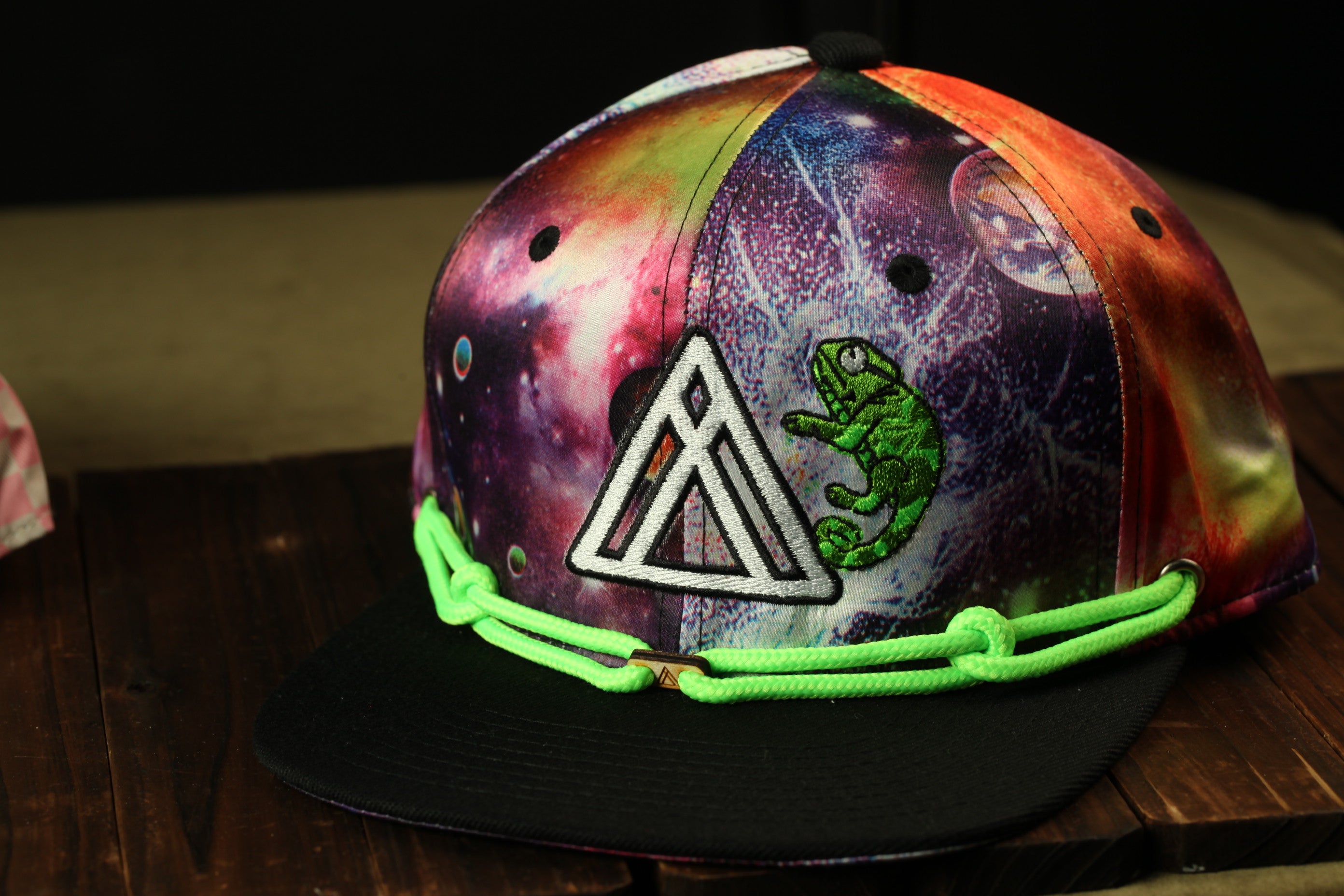 A colorful, patterned baseball cap with a space-themed design, featuring a green chameleon and a triangular logo.