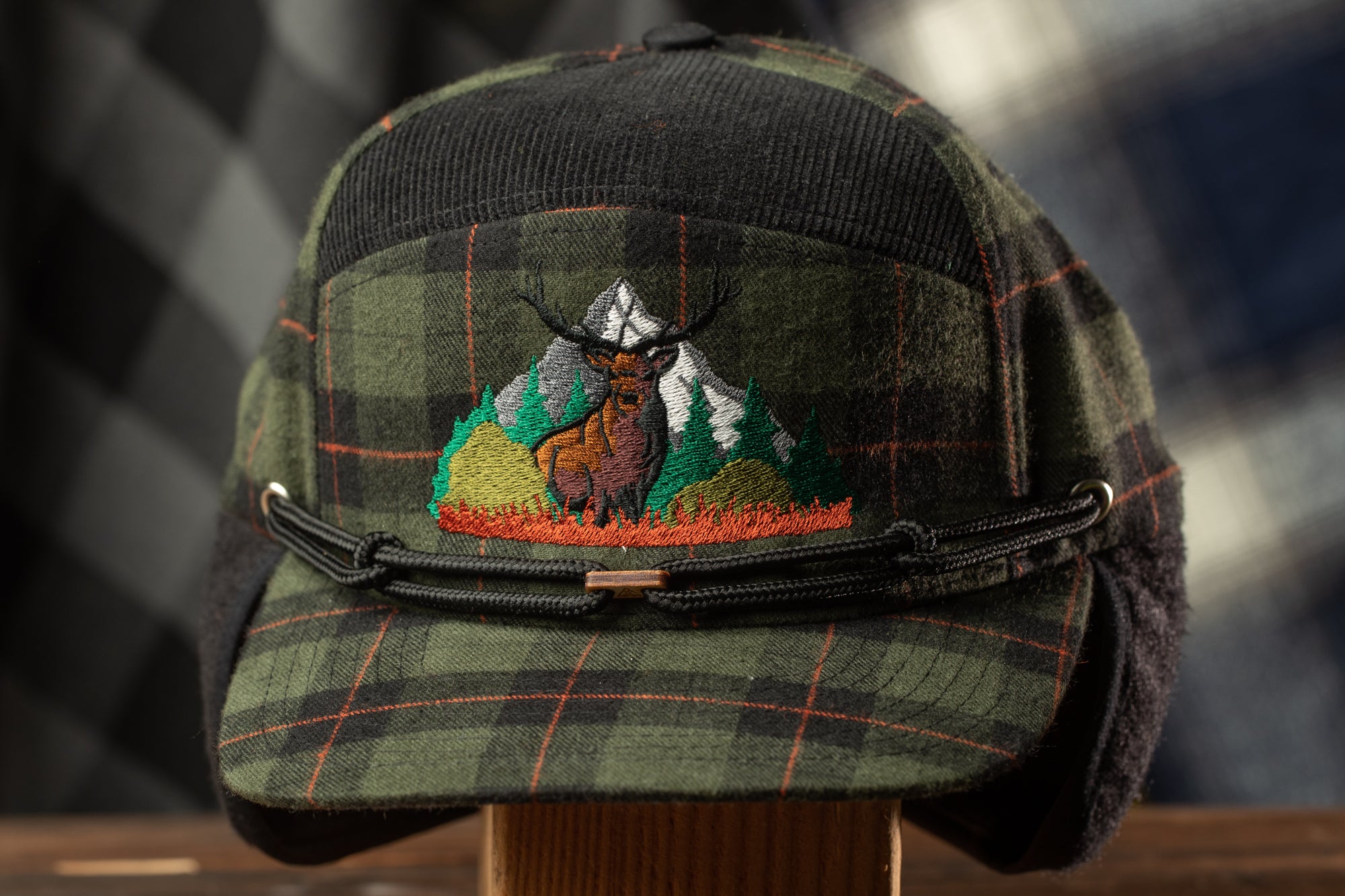 A plaid hat with a mountain and deer embroidered on it, displayed on a wooden stand.