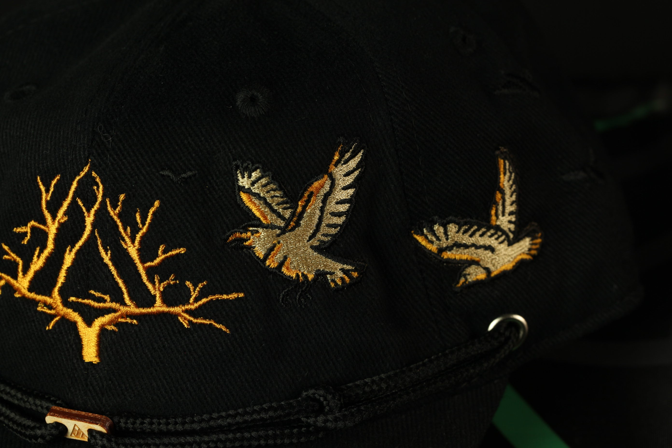 a close-up view of a black baseball cap with gold embroidered designs. The cap features a branch with golden leaves and two birds in flight, one on each side.
