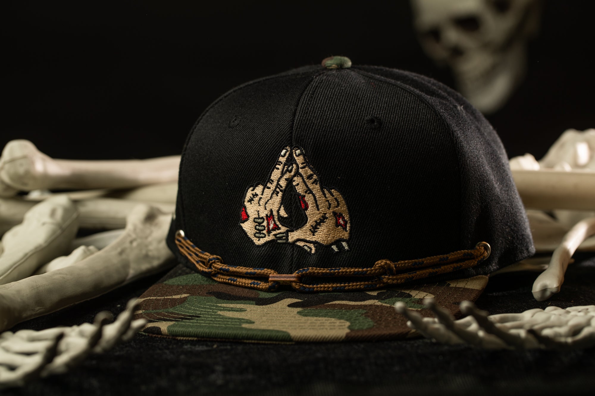 A black baseball cap with a camouflage pattern and a skull design on it, placed on a surface with bones and a skull.