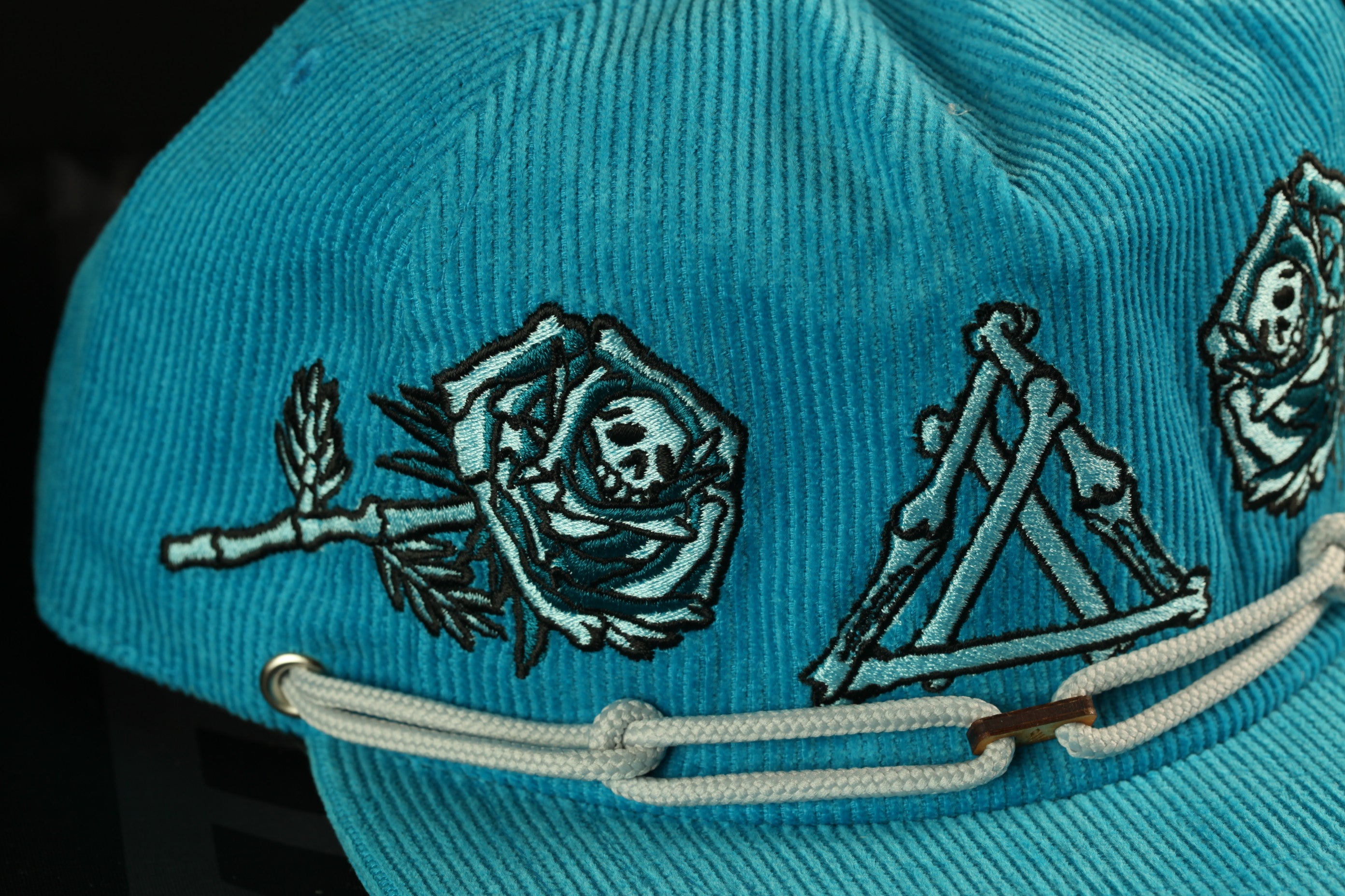 A close-up view of a teal-colored baseball cap with various embroidered designs, including a rose, skeleton, and triangle motifs.