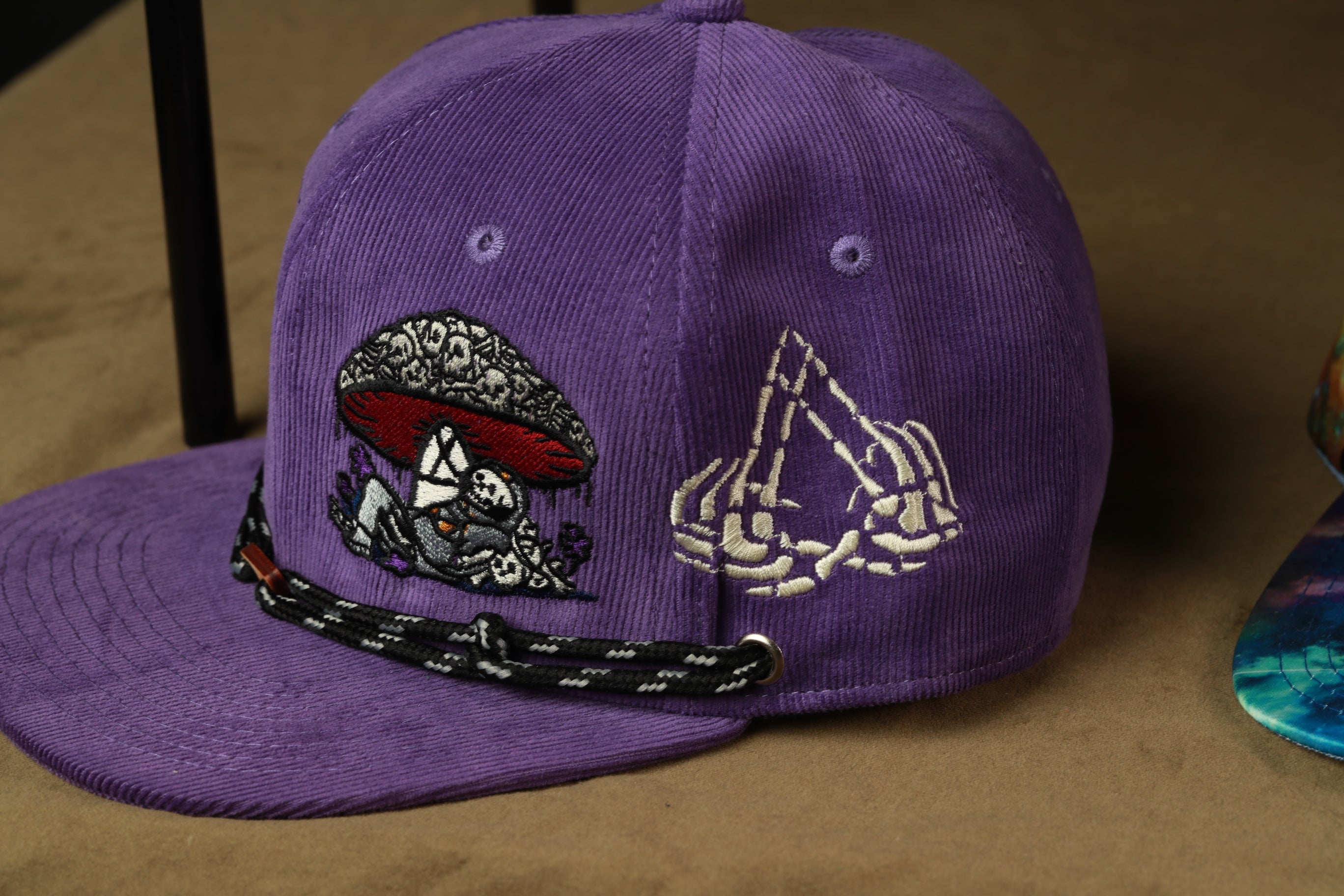 A purple baseball cap with various embroidered designs, including a mushroom, skeleton hands, and a black and white striped strap.