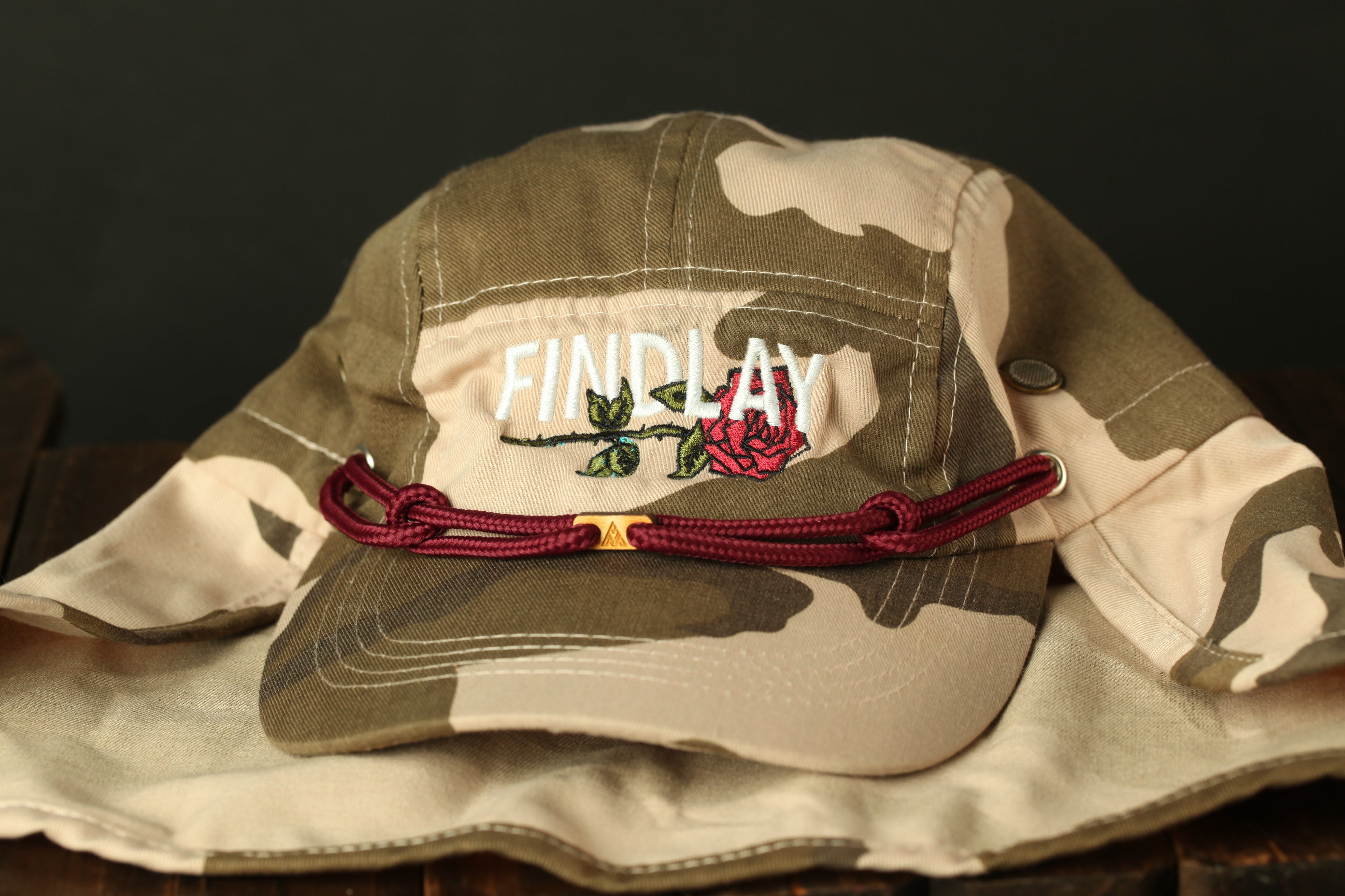 A camouflage-patterned baseball cap with the word "FINDlay" printed on it, along with a red string or strap.