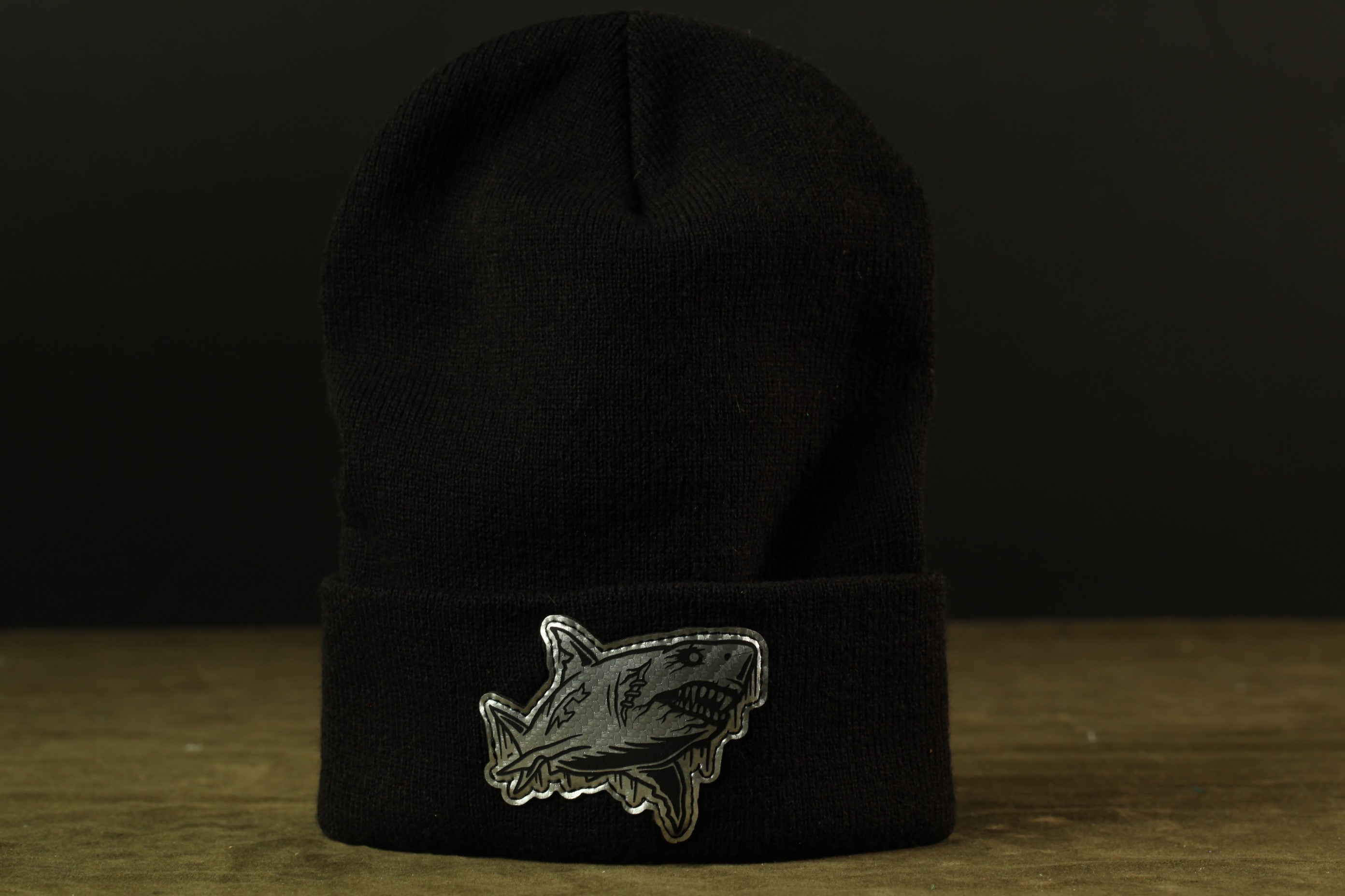 A black knit beanie with a shark patch on the front is displayed against a dark background.