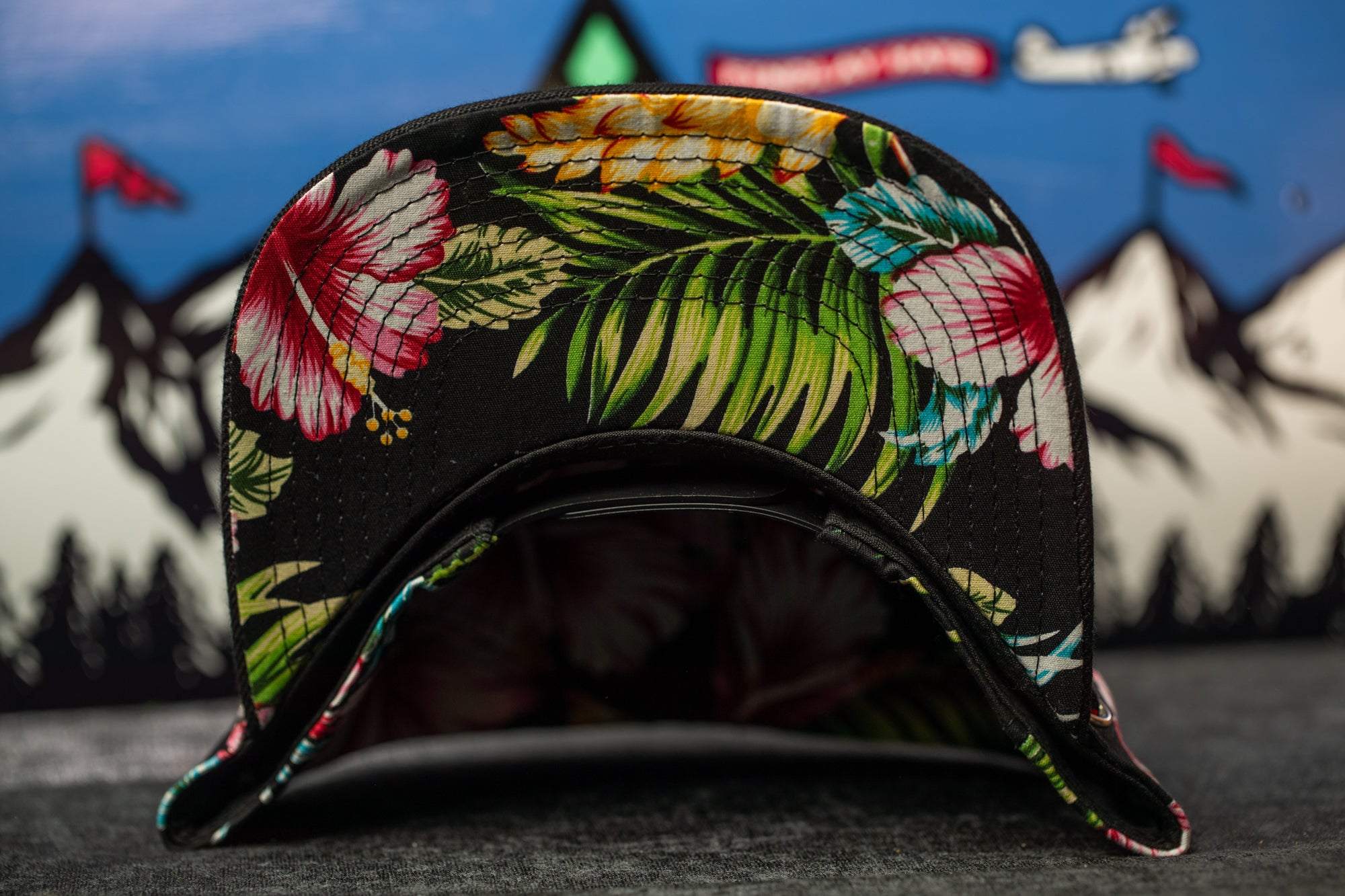 A close-up view of a colorful floral-patterned baseball cap, with a blurred background featuring mountains and flags.
