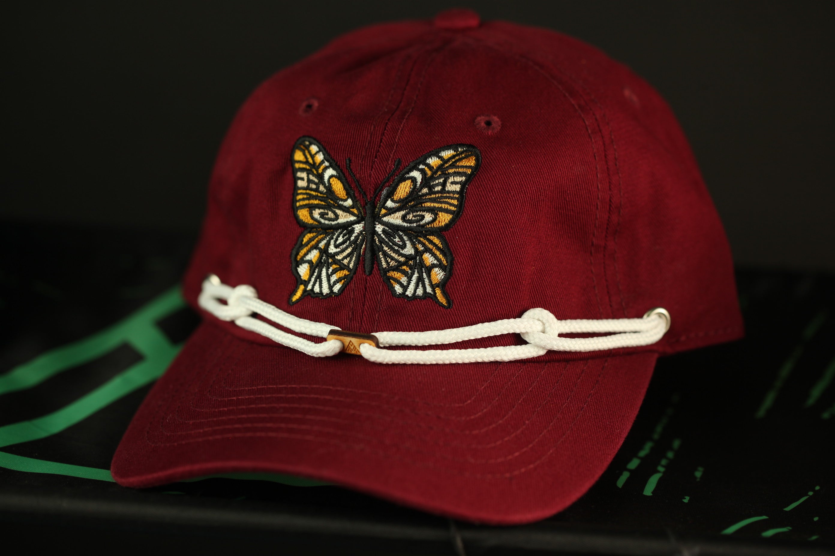 A maroon baseball cap with a white butterfly embroidered on it, resting on a black surface.