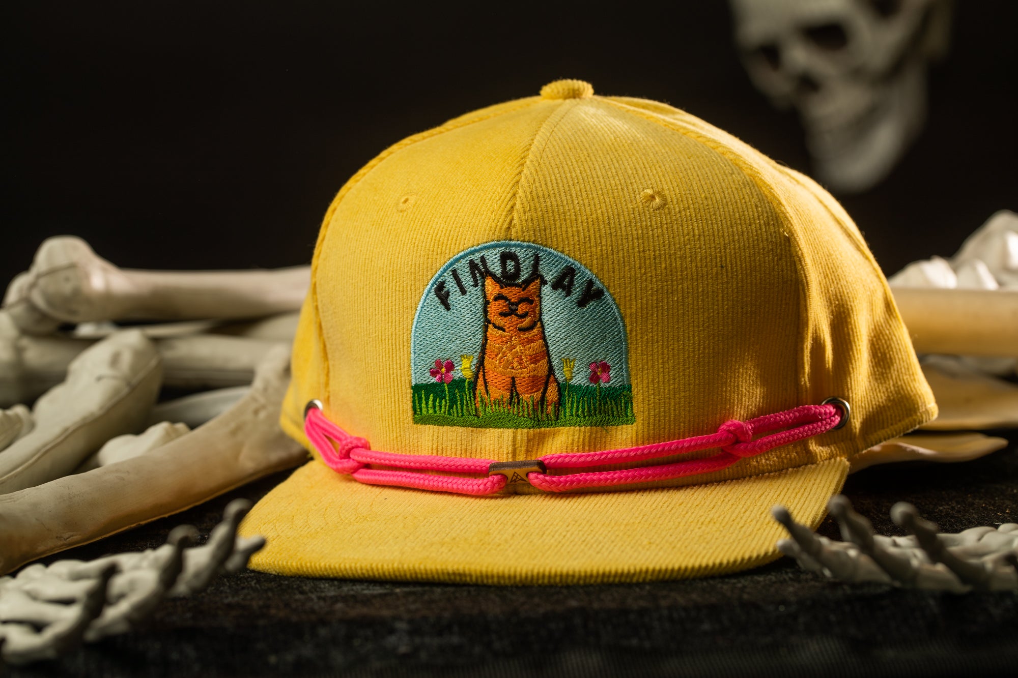 A yellow baseball cap with a cartoon image of a cat and the word "FINDAY" on it, along with a pink string around the brim.