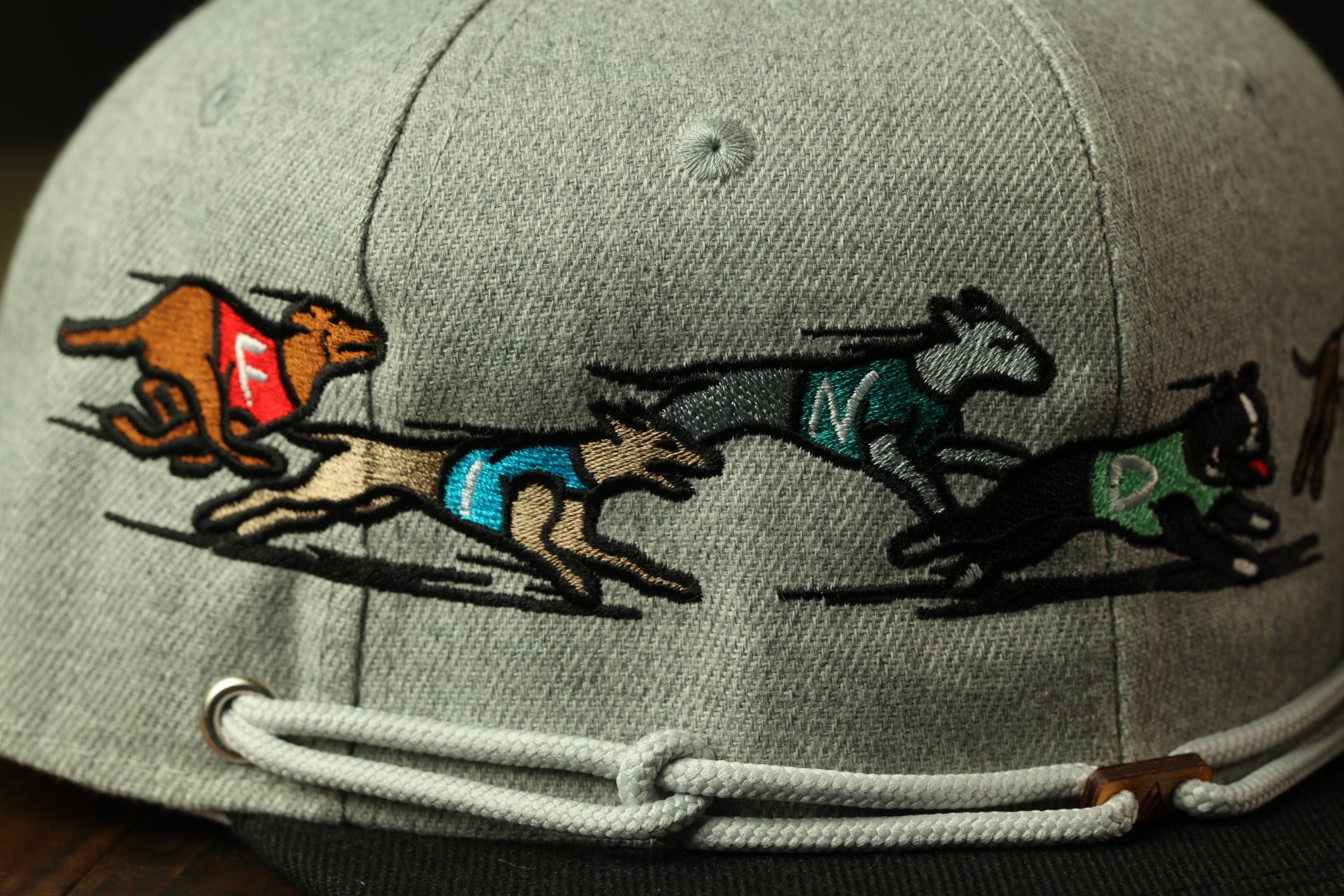 A close-up view of a gray baseball cap with a detailed embroidered design featuring three dogs running at high speeds.