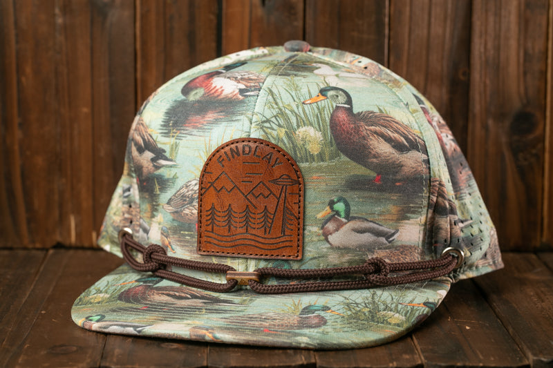 A camouflage baseball cap with a printed design of ducks and other wildlife, including a leather patch with a mountain landscape.