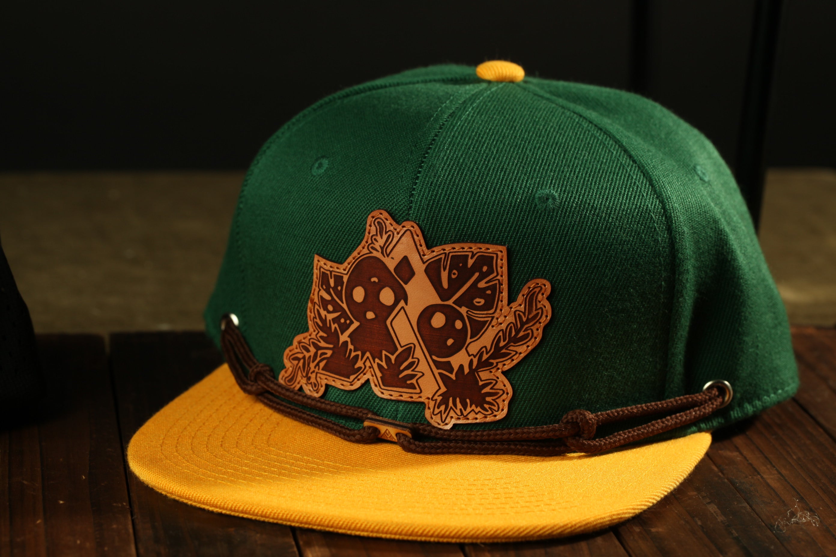 A green baseball cap with a brown leather patch featuring a leaf design and a yellow brim, placed on a wooden surface.