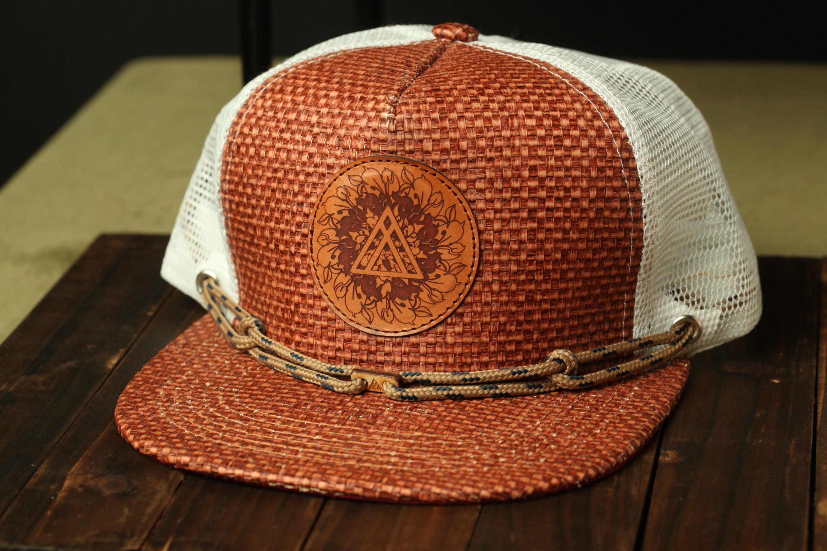 A brown woven baseball cap with a white mesh back, featuring a circular logo on the front.